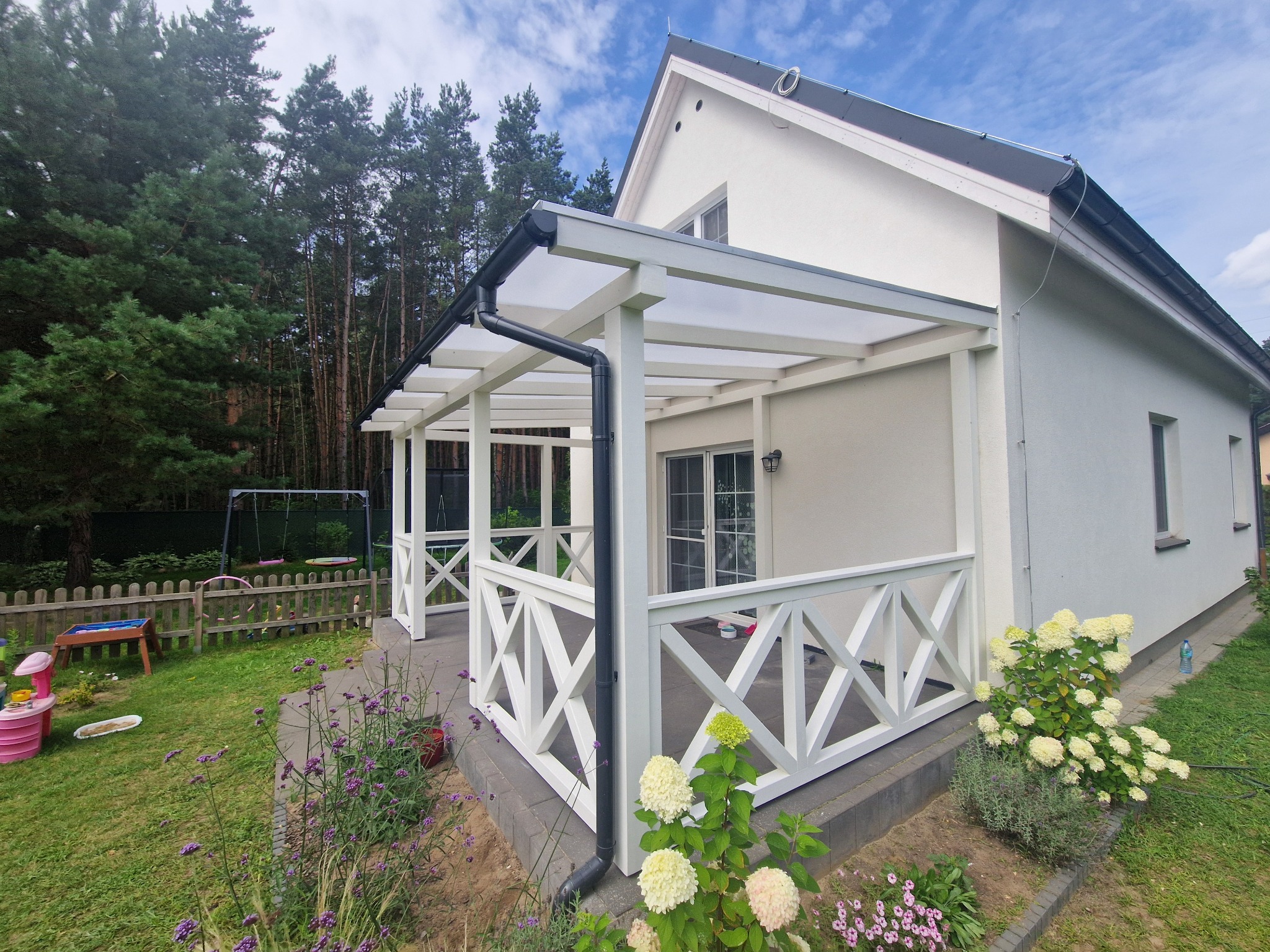 Biała pergola z przezroczystym dachem przylegająca do jasnego domu, z dekoracyjną balustradą i widokiem na ogród z kwiatami.