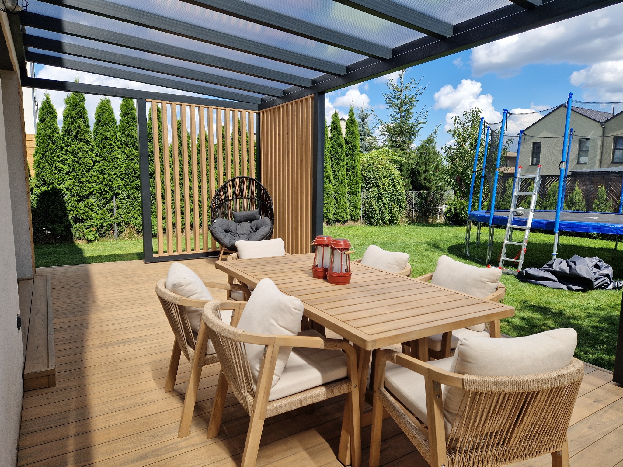 Zadaszony taras z drewnianą podłogą, stołem i krzesłami w stylu boho. W tle zielony ogród z trampoliną i tujami. Widoczna ażurowa ścianka i wiszące krzesło.