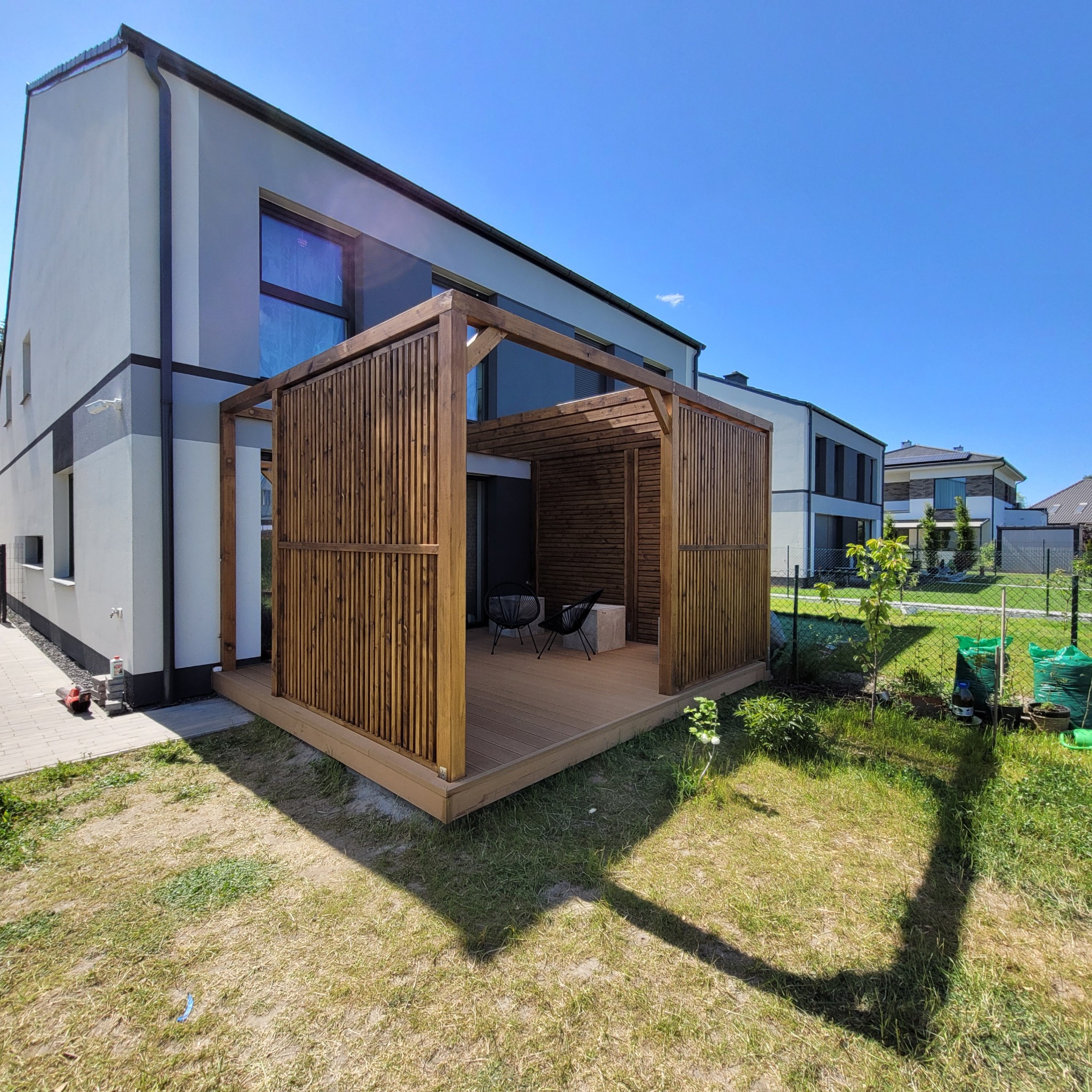 Nowoczesna, drewniana konstrukcja z ażurowymi ścianami, zlokalizowana na tarasie z desek kompozytowych, z dwoma krzesłami wewnątrz, na tle nowoczesnego domu i błękitnego nieba.