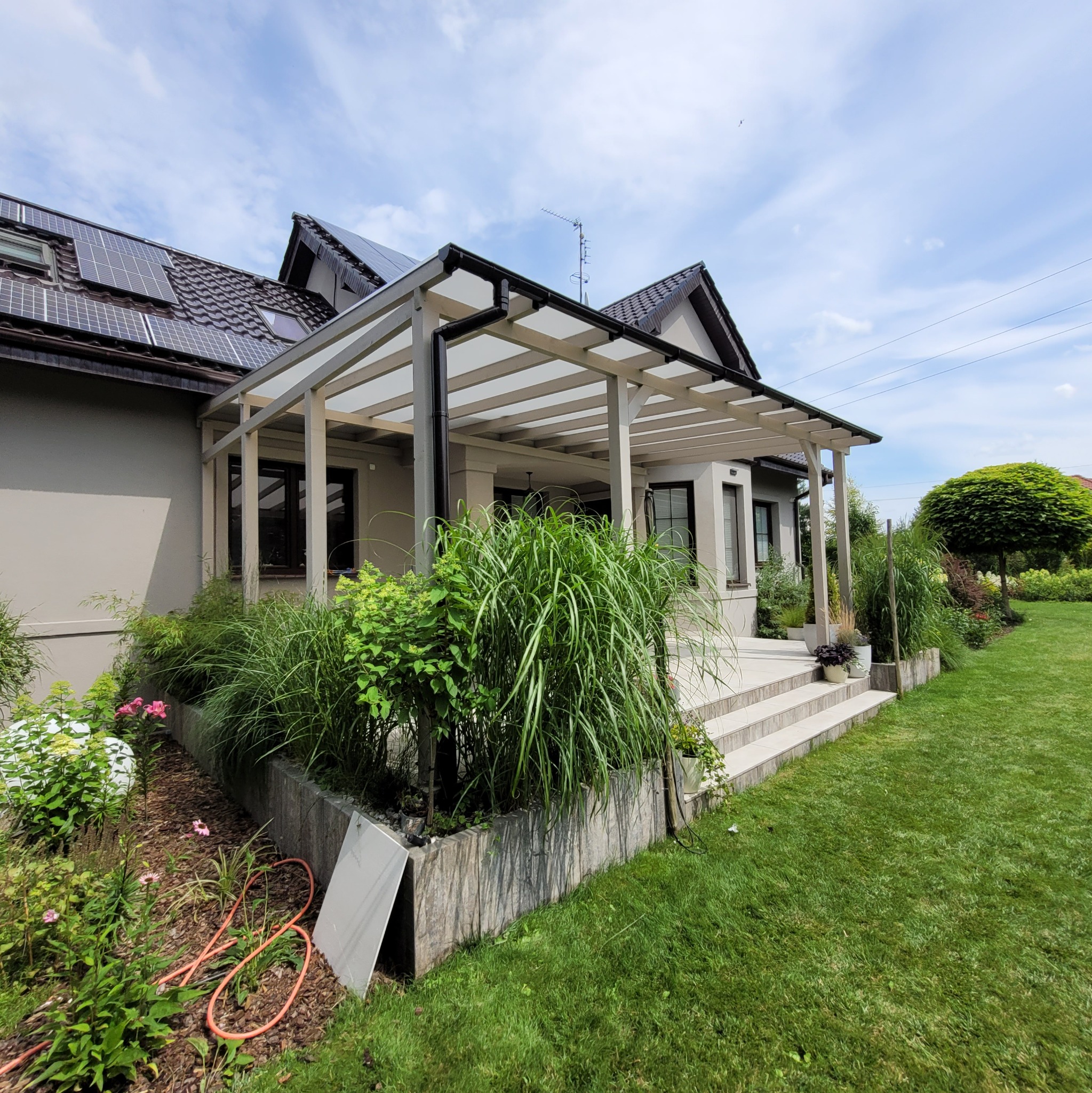 Zadaszony taras z jasną konstrukcją drewnianą, schodami z płytek i bujną roślinnością w donicach oraz wzdłuż podwyższonej rabaty kwiatowej, widok na dom z panelami słonecznymi.