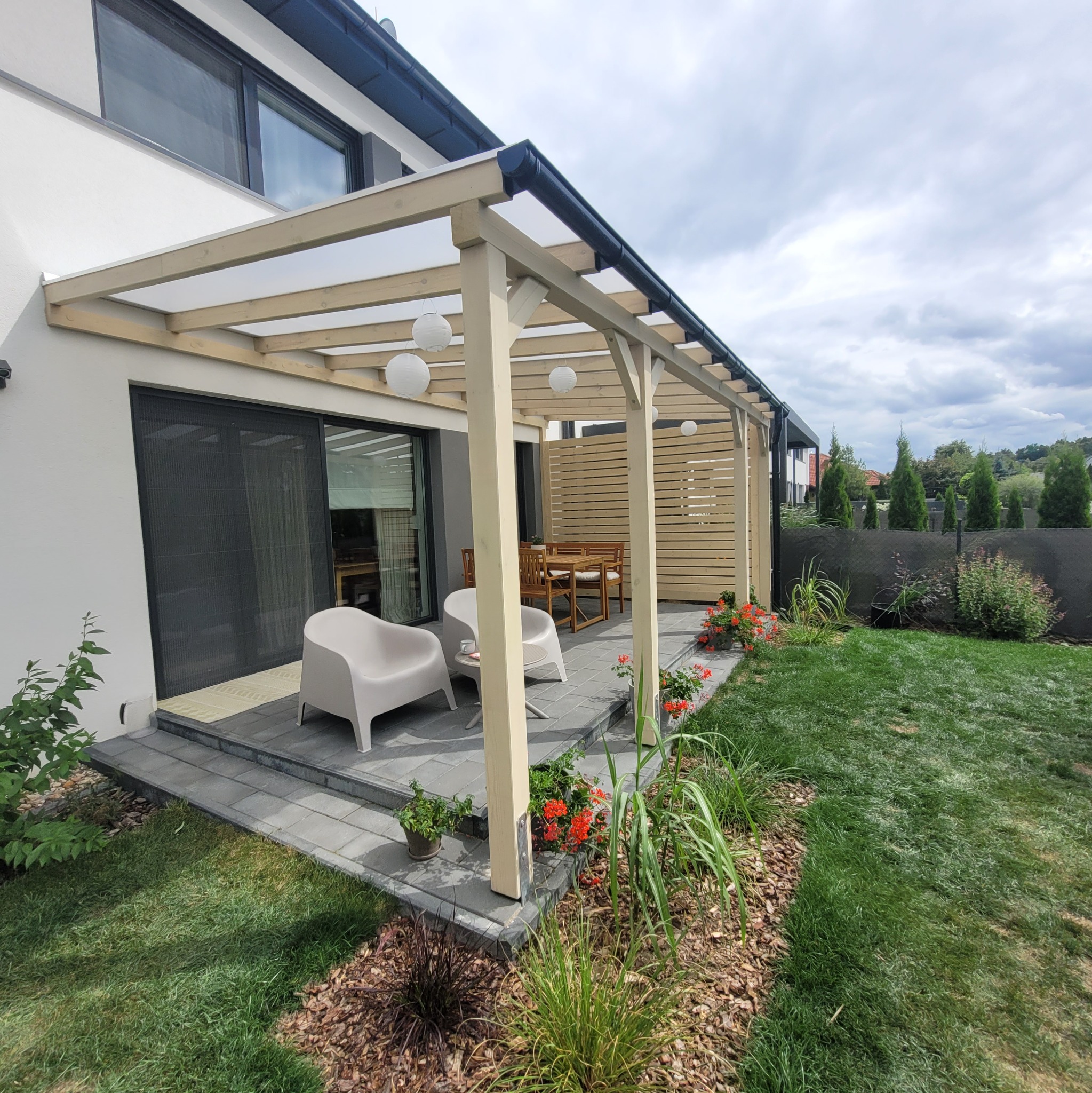 Nowoczesna pergola z drewnianą konstrukcją i przezroczystym zadaszeniem, oświetlona kulistymi lampami, przylegająca do białego domu z szarym tarasem i meblami ogrodowymi, otoczona zielonym...