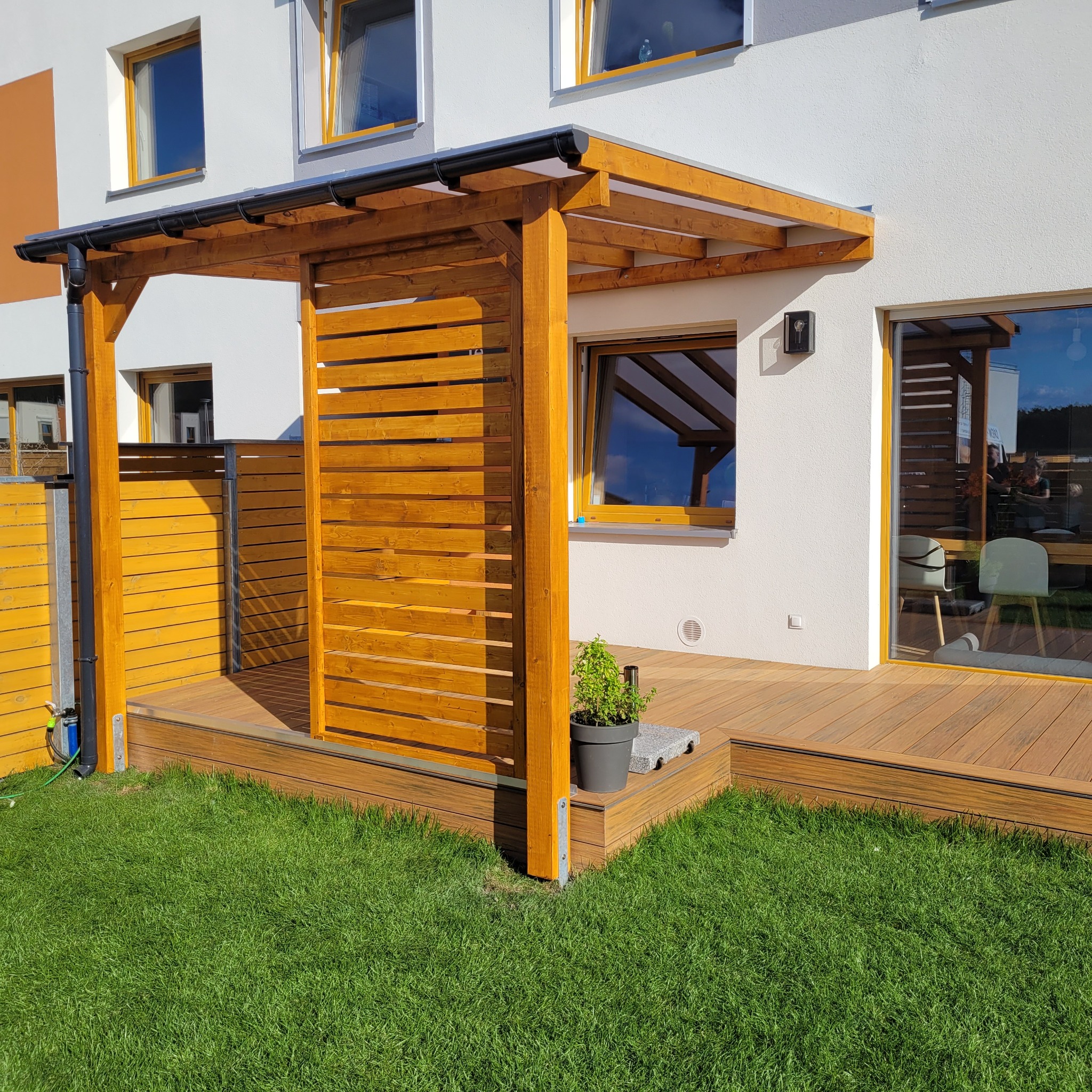 Nowoczesna konstrukcja z drewna zadaszająca taras, z elementami ażurowymi i przezroczystym dachem, połączona z drewnianym tarasem kompozytowym przylegającym do białego budynku z dużymi oknami,...