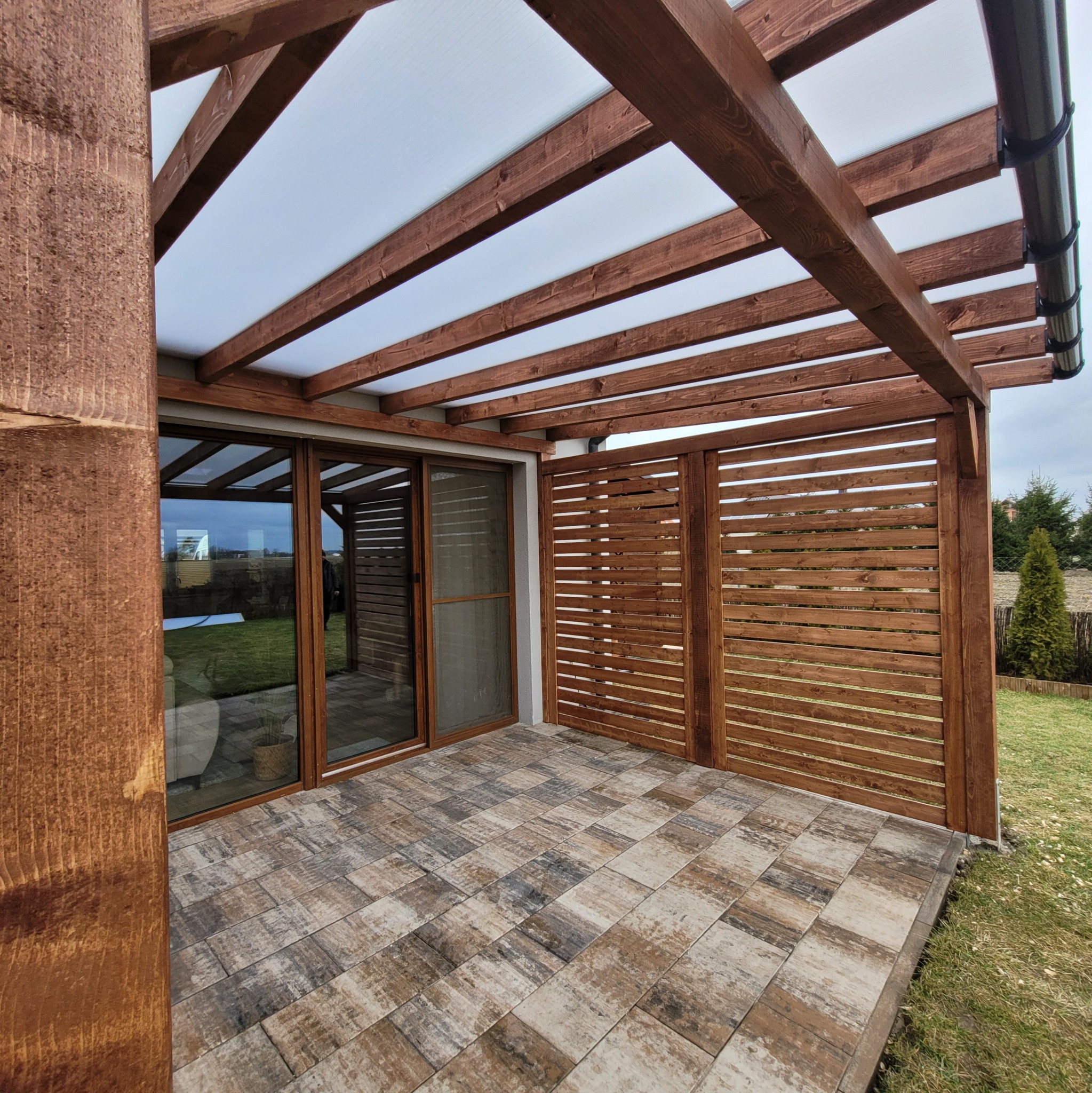 Zadaszona konstrukcja z drewnianą pergolą i ażurową ścianką w Poznaniu, przylegająca do budynku z przesuwnymi drzwiami, wyłożona kostką brukową w odcieniach brązu i szarości.