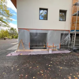 Przygotowania do malowania elewacji domu: zabezpieczone okna i ściany folią ochronną, drabina oparta o ścianę, mały stolik z narzędziami i farbami, widoczne rusztowanie z boku budynku.