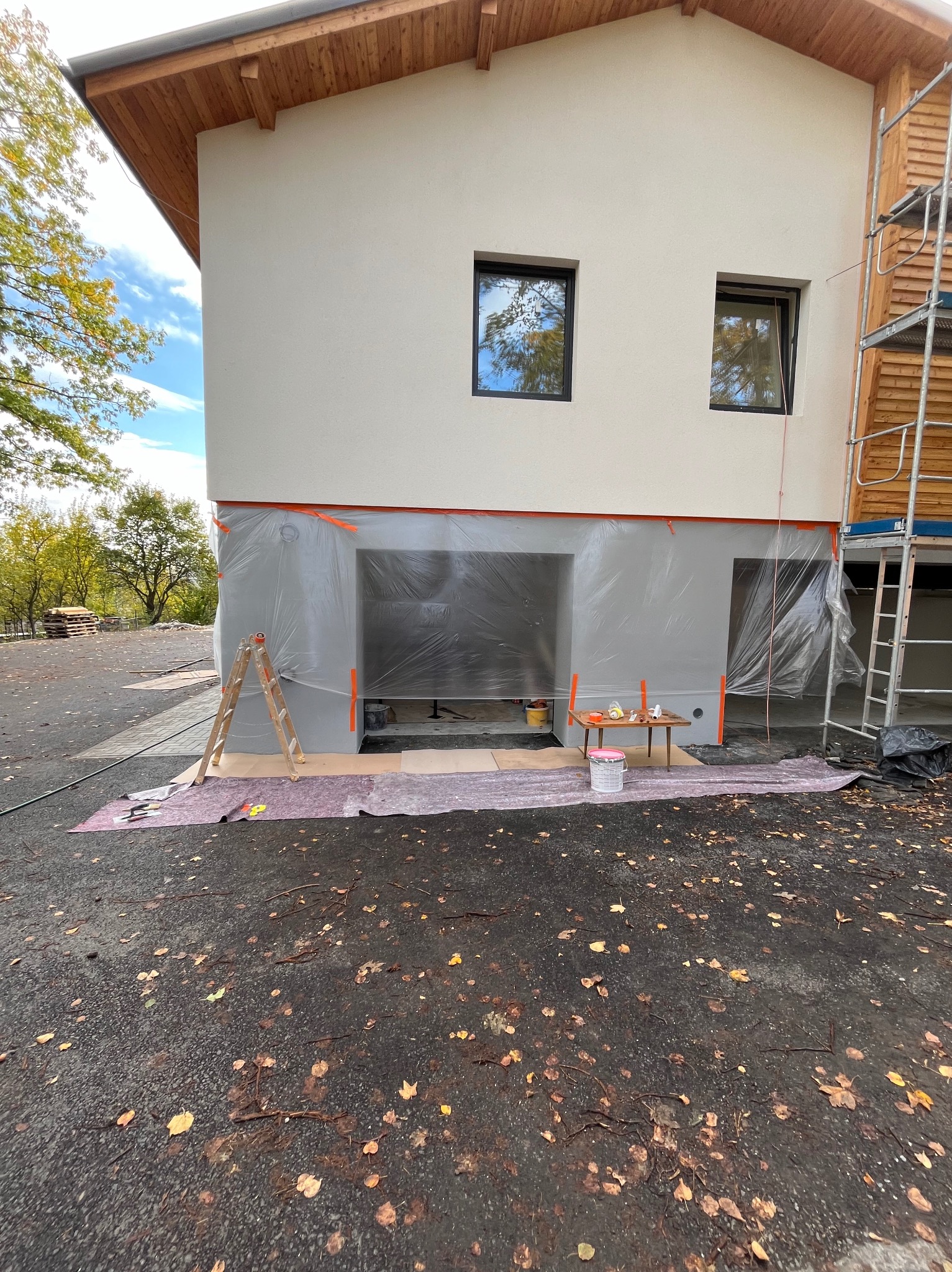 Przygotowania do malowania elewacji domu: zabezpieczone okna i ściany folią ochronną, drabina oparta o ścianę, mały stolik z narzędziami i farbami, widoczne rusztowanie z boku budynku.