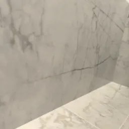 Ściana i podłoga wyłożone białymi płytkami imitującymi marmur z delikatnymi szarymi i brązowymi żyłkami, widoczne fugi.