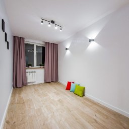 SAVCHUK-BUD SPÓŁKA Z OGRANICZONĄ ODPOWIEDZIALNOŚCIĄ - Jasne wnętrze pokoju po remoncie z drewnianą podłogą, trzema kolorowymi poduszkami przy ścianie, grzejnikiem pod oknem z różowymi zasłonami i minimalistycznym oświetleniem sufitowym i ściennym.