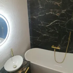 Nowoczesna łazienka z białymi, wzorzystymi płytkami, czarnymi płytkami imitującymi marmur, białą owalną umywalką nablatową na czarnej szafce z elementami złota oraz białą wanną z baterią w kolorze...