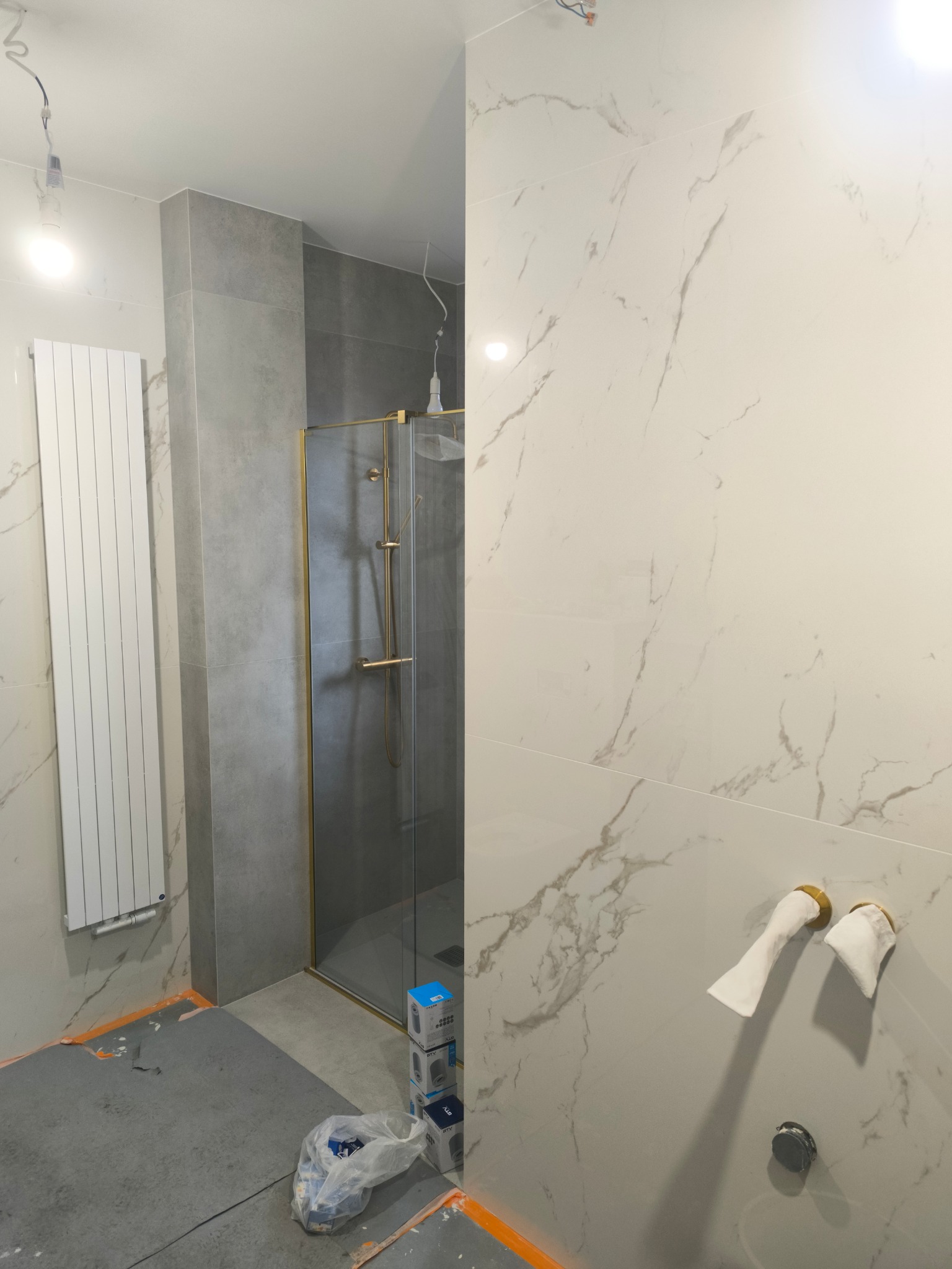 Fragment łazienki w trakcie wykańczania: białe płytki imitujące marmur, złota rama kabiny prysznicowej, grzejnik, zabezpieczone rury, podłoga zabezpieczona folią.