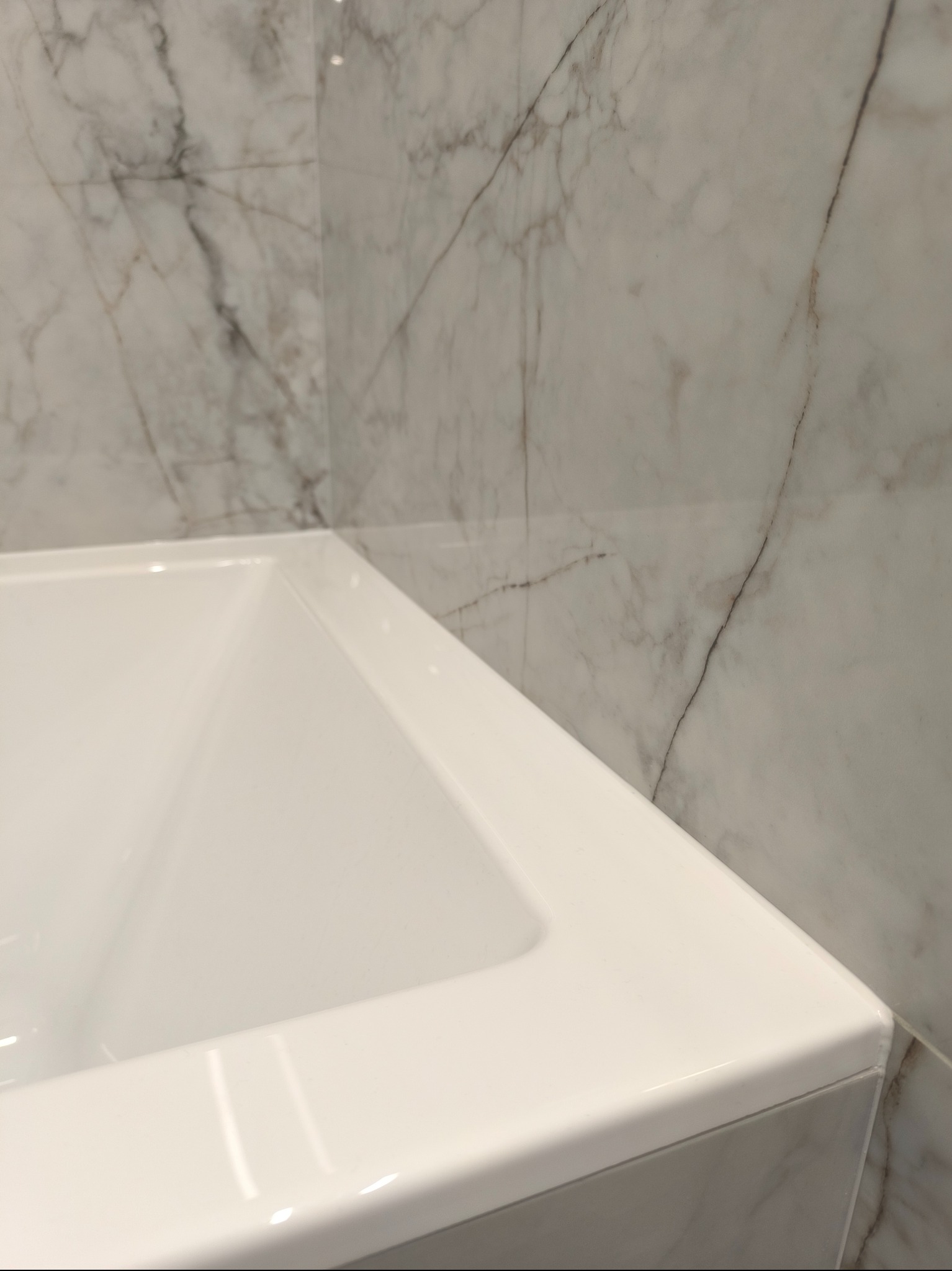 Biała wanna narożna, z widocznymi szarymi płytkami imitującymi marmur na ścianach, z delikatnymi refleksami światła.