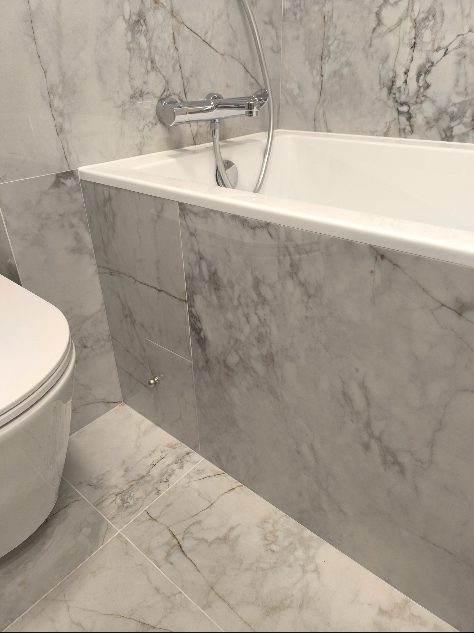 Fragment łazienki z białą wanną, toaletą i marmurowymi płytkami na ścianach i podłodze, z widocznym chromowanym kranem i słuchawką prysznicową.