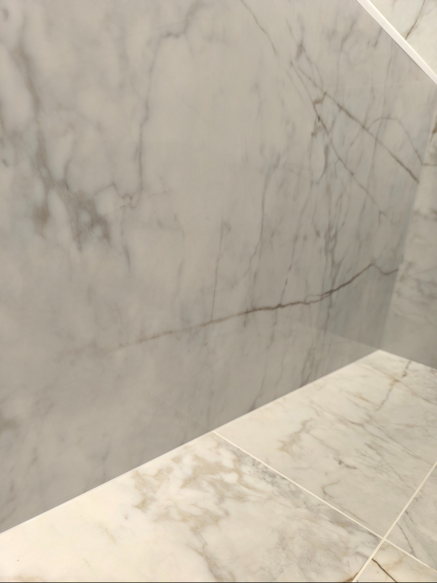 Ściana i podłoga wyłożone białymi płytkami imitującymi marmur z delikatnymi szarymi i brązowymi żyłkami, widoczne fugi.