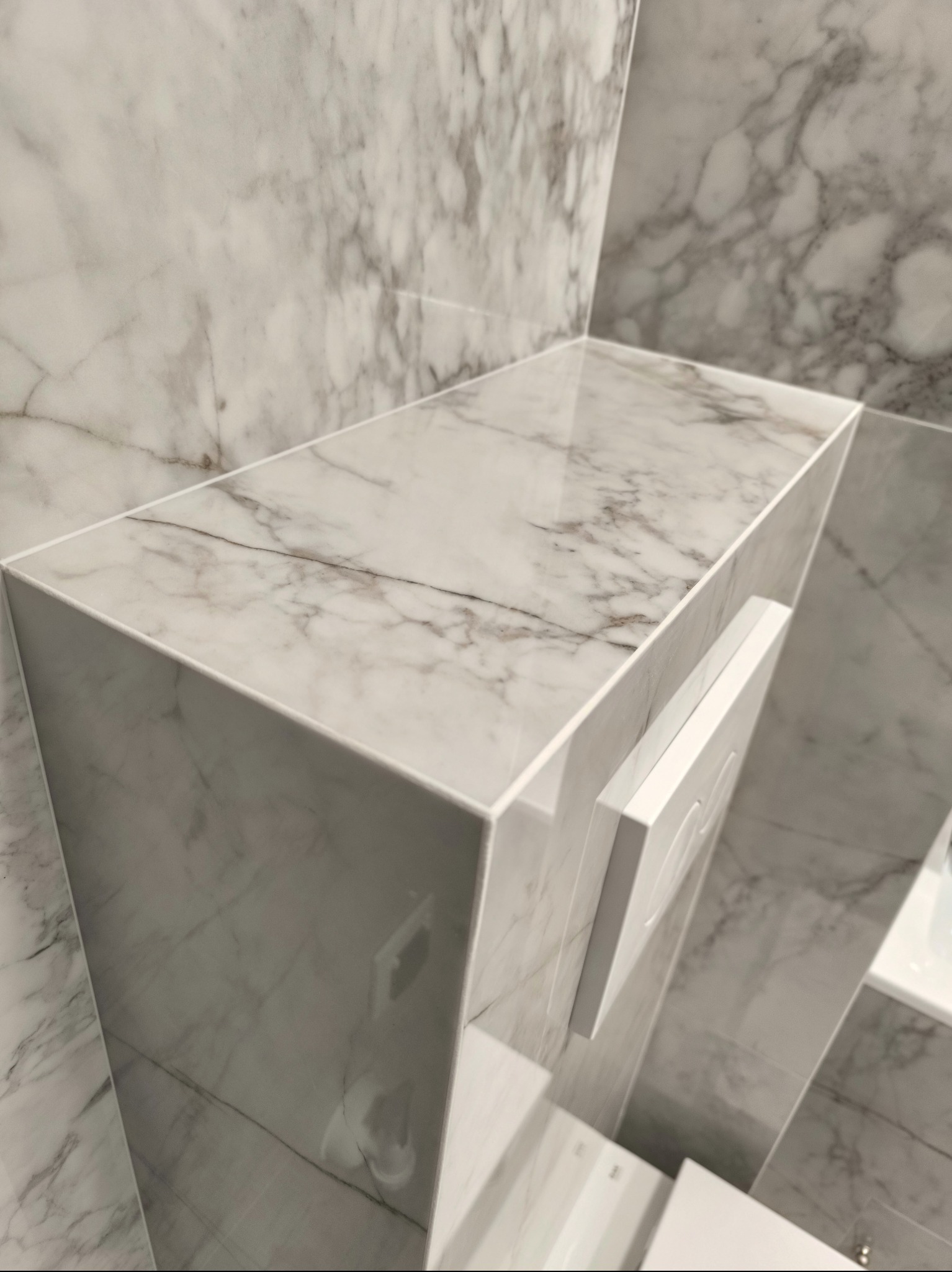 Półka i zabudowa stelaża WC wykończone płytkami imitującymi marmur Calacatta, widoczny biały przycisk spłuczki.