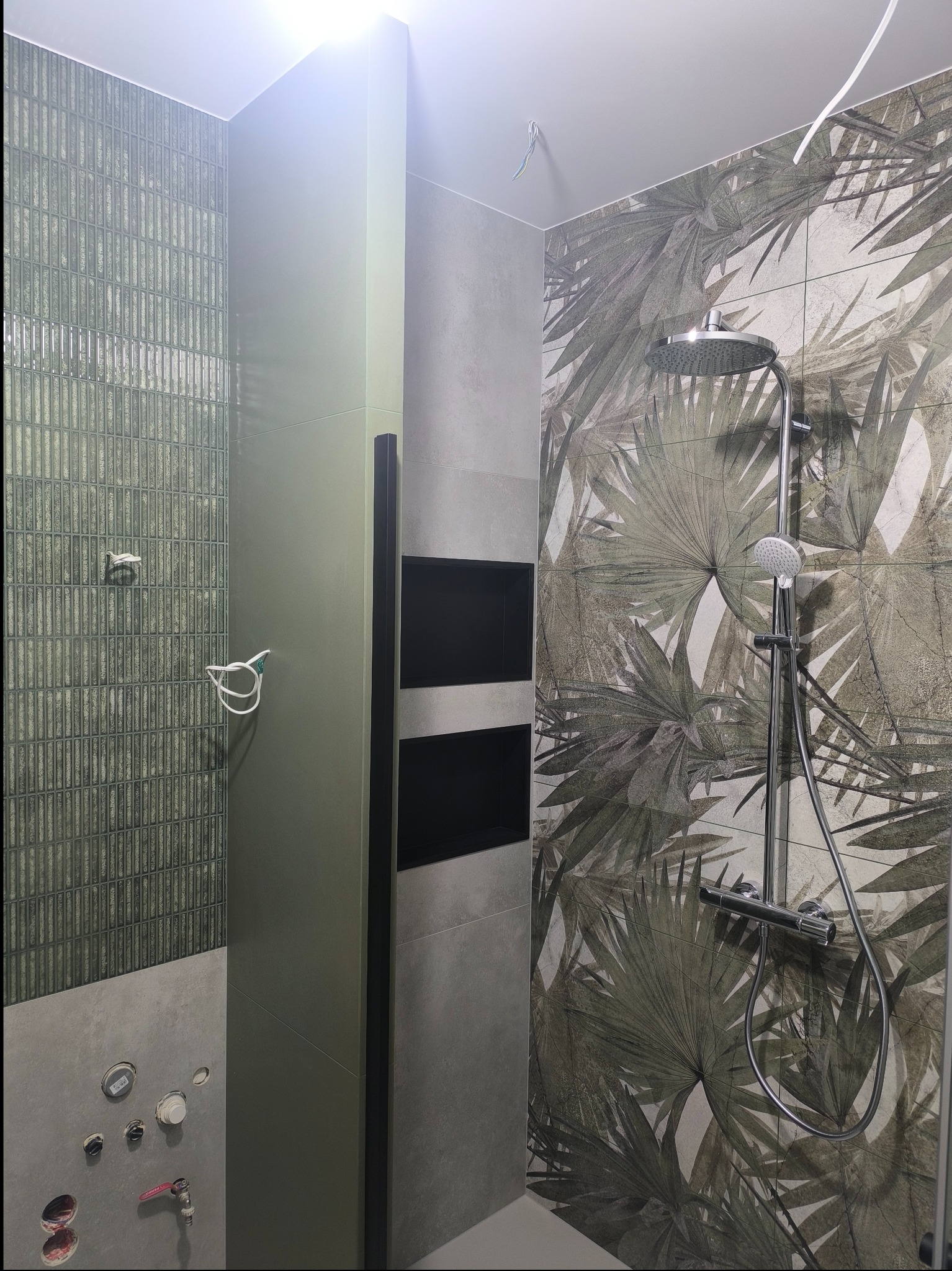 Wnętrze łazienki w trakcie wykańczania: widoczne płytki w odcieniach zieleni z motywem roślinnym, kabina prysznicowa z deszczownicą, oraz przygotowane punkty instalacyjne na ścianie.