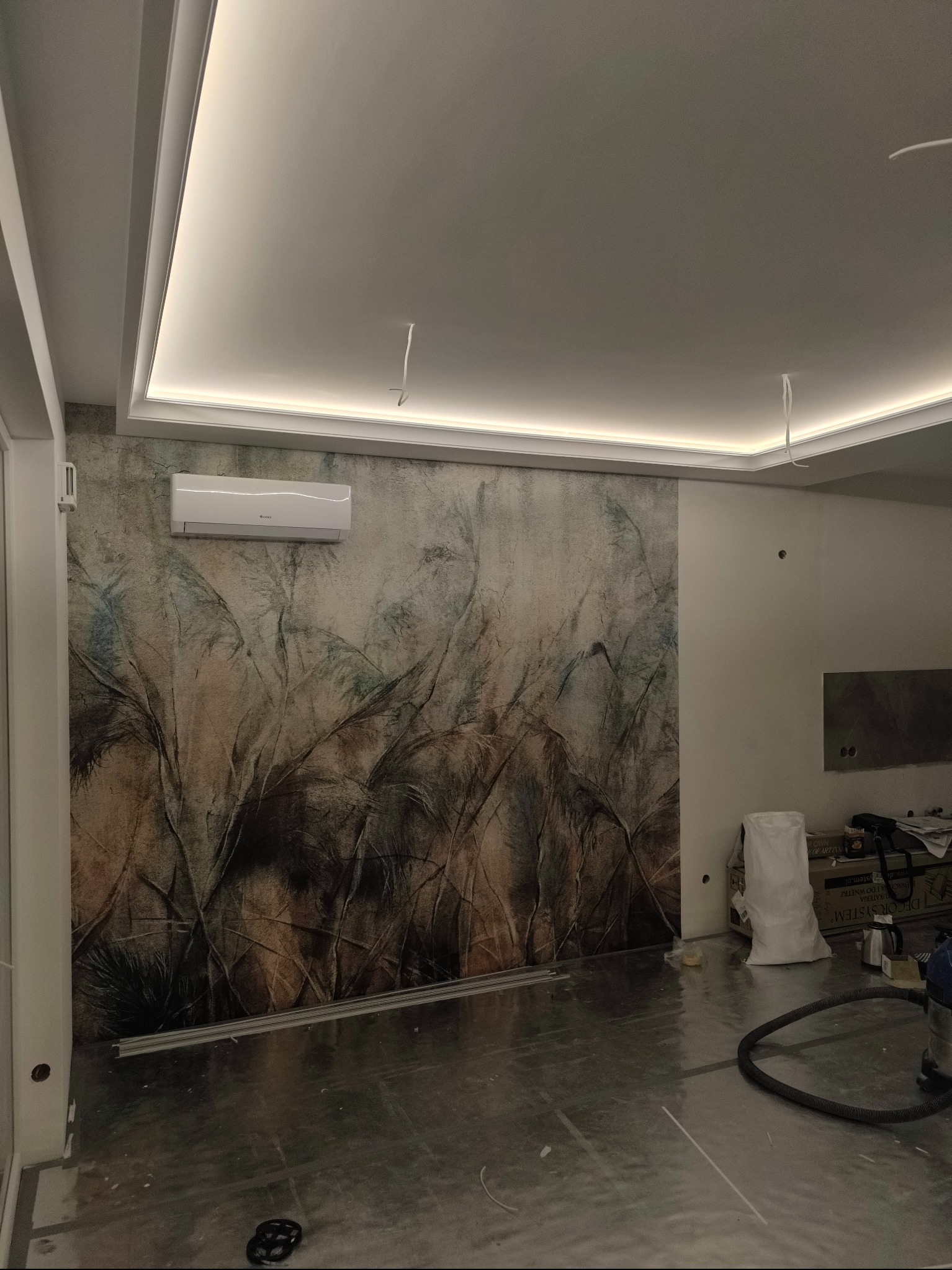 Pokój w trakcie wykańczania, z dekoracyjną tapetą z motywem roślinnym, klimatyzacją na ścianie, podwieszanym sufitem z oświetleniem LED, widoczne przewody elektryczne oraz materiały budowlane.