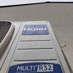 Mart-Klim Marta Pełczyńska - Zewnętrzna jednostka klimatyzacji Haier Multi R32 Inverter, pokryta kroplami wody, zamontowana na szarej elewacji budynku, widok z dołu.