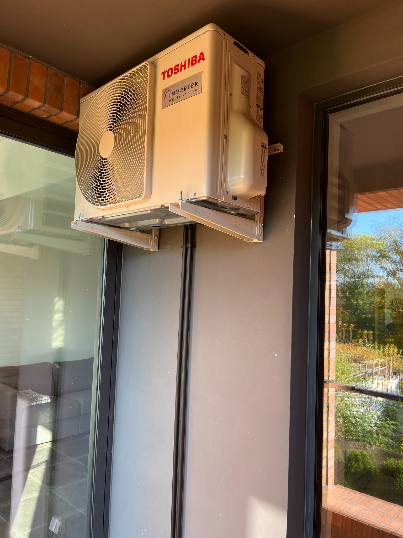 Zewnętrzna jednostka klimatyzacji Toshiba Inverter zamontowana na ścianie budynku, widok z boku, z widocznym fragmentem balkonu i zieleni za oknem.