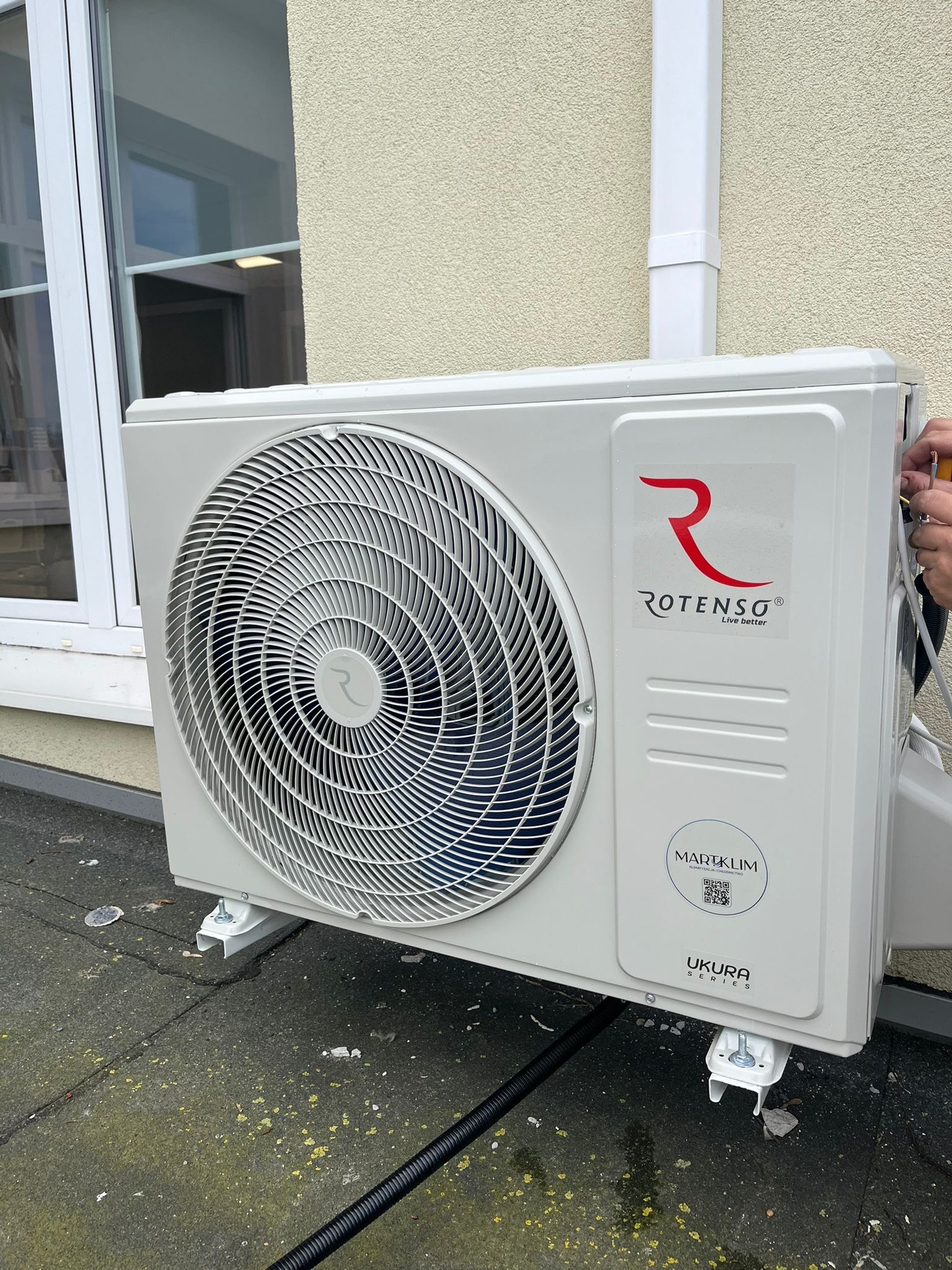 Zewnętrzna jednostka klimatyzacji Rotenso zamontowana na zewnątrz budynku, widoczny kabel i fragment okna.