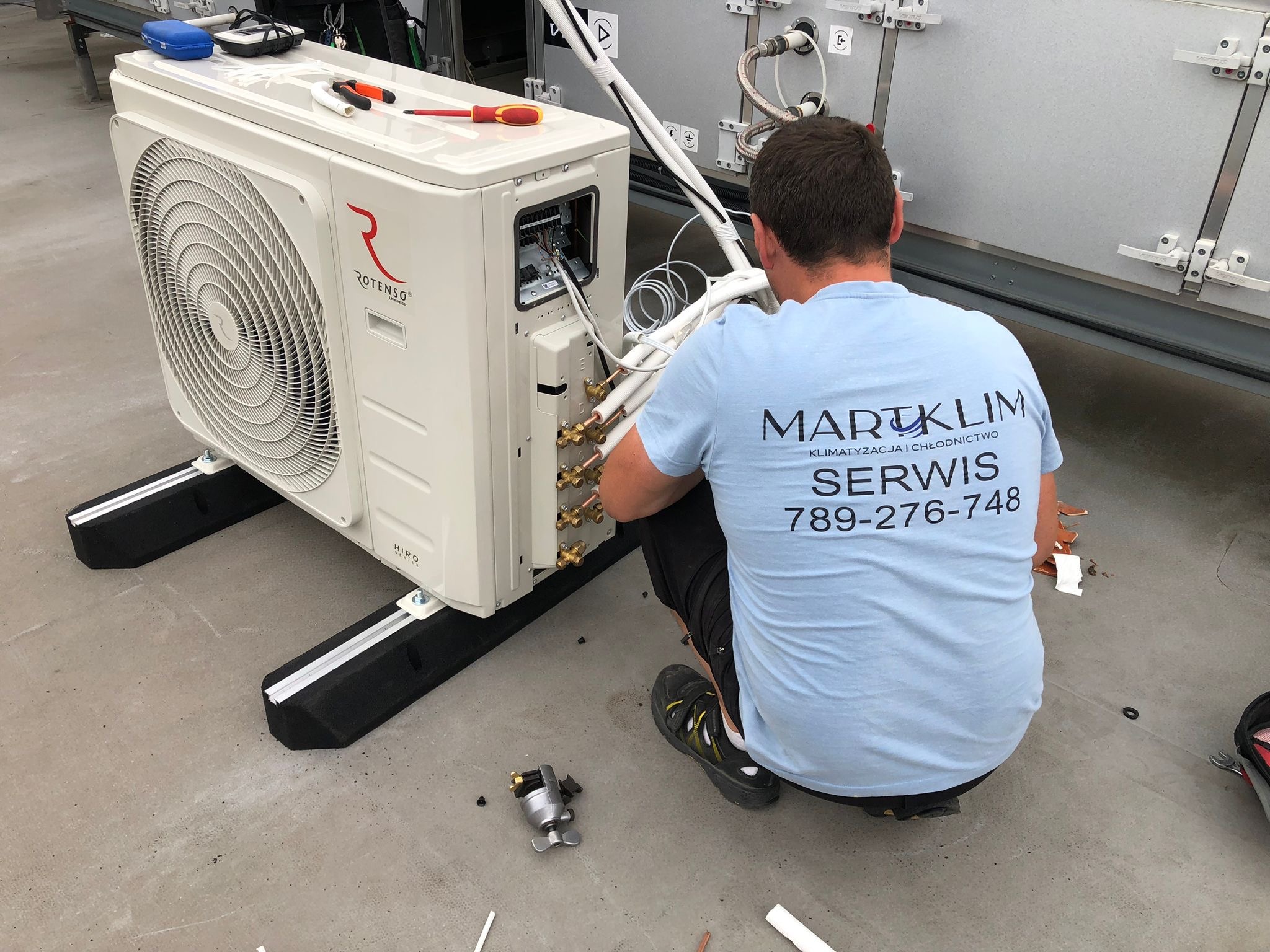 Instalator w koszulce firmowej 'MARTKLIM' podłącza przewody do zewnętrznej jednostki klimatyzatora Rotenso na dachu. Widoczne narzędzia, kable i elementy montażowe.