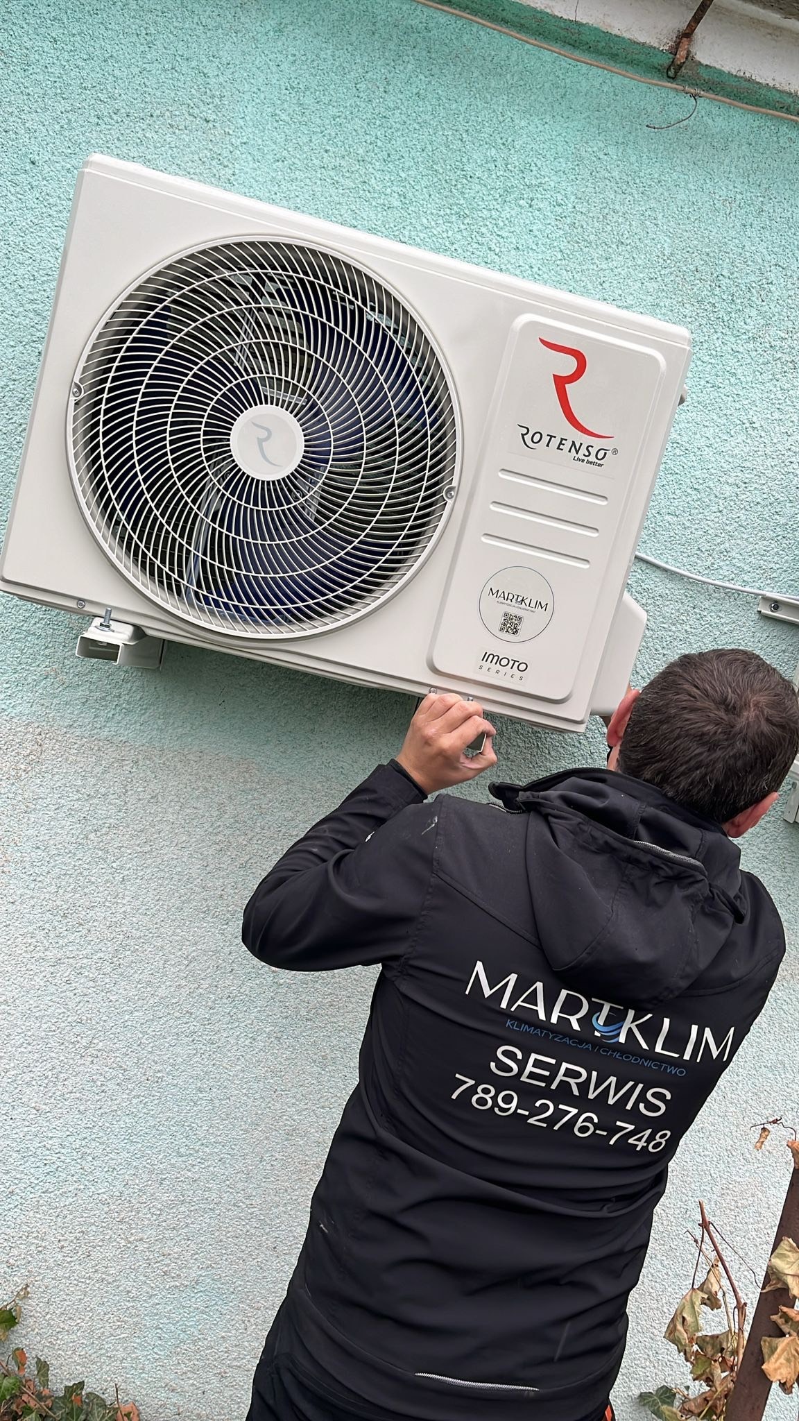Instalacja jednostki zewnętrznej klimatyzacji Rotenso w Głuchowie, widoczny monter w kurtce firmowej Martklim, przytwierdzający urządzenie do ściany o szorstkiej fakturze.