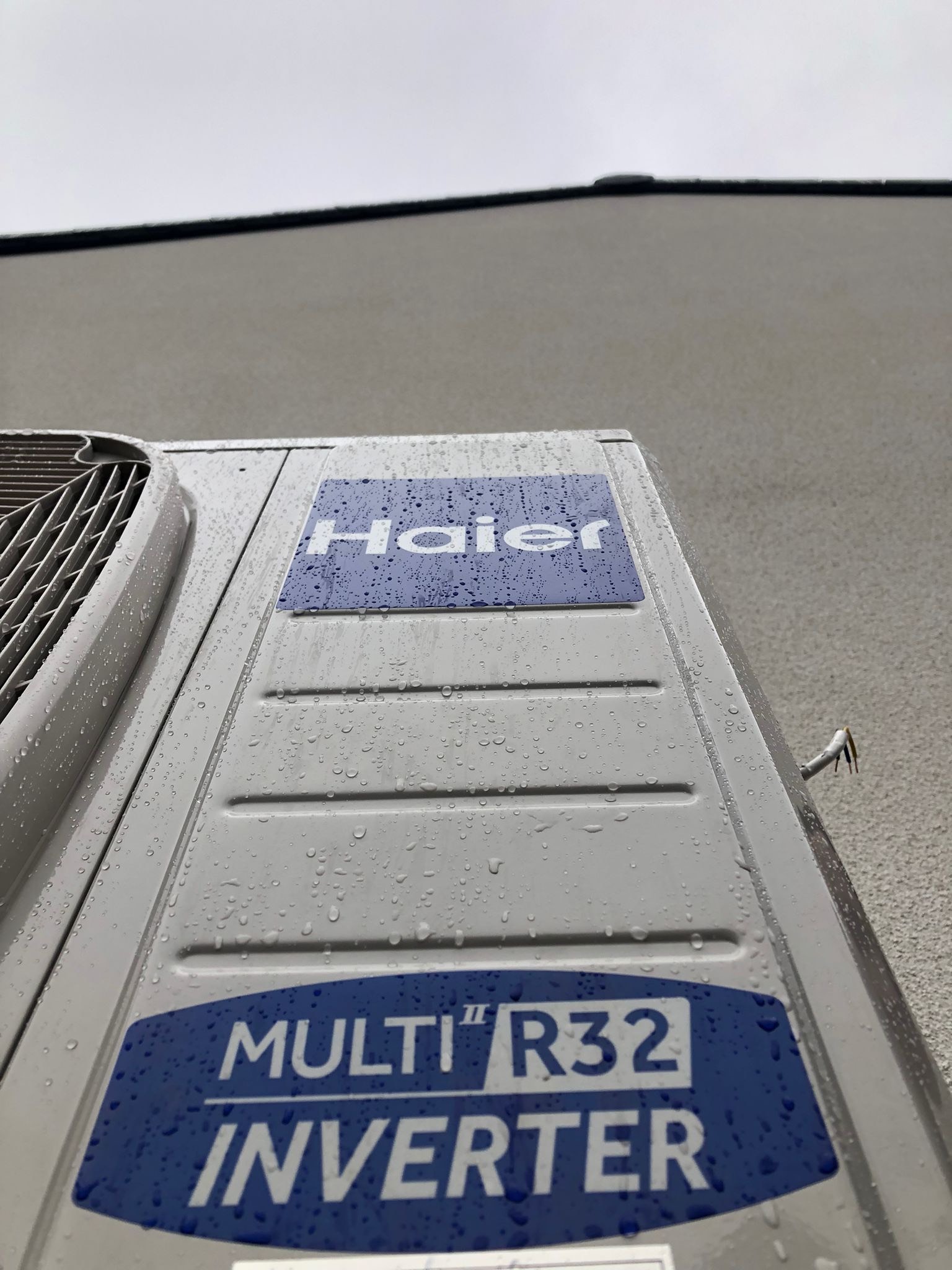 Zewnętrzna jednostka klimatyzacji Haier Multi R32 Inverter, pokryta kroplami wody, zamontowana na szarej elewacji budynku, widok z dołu.
