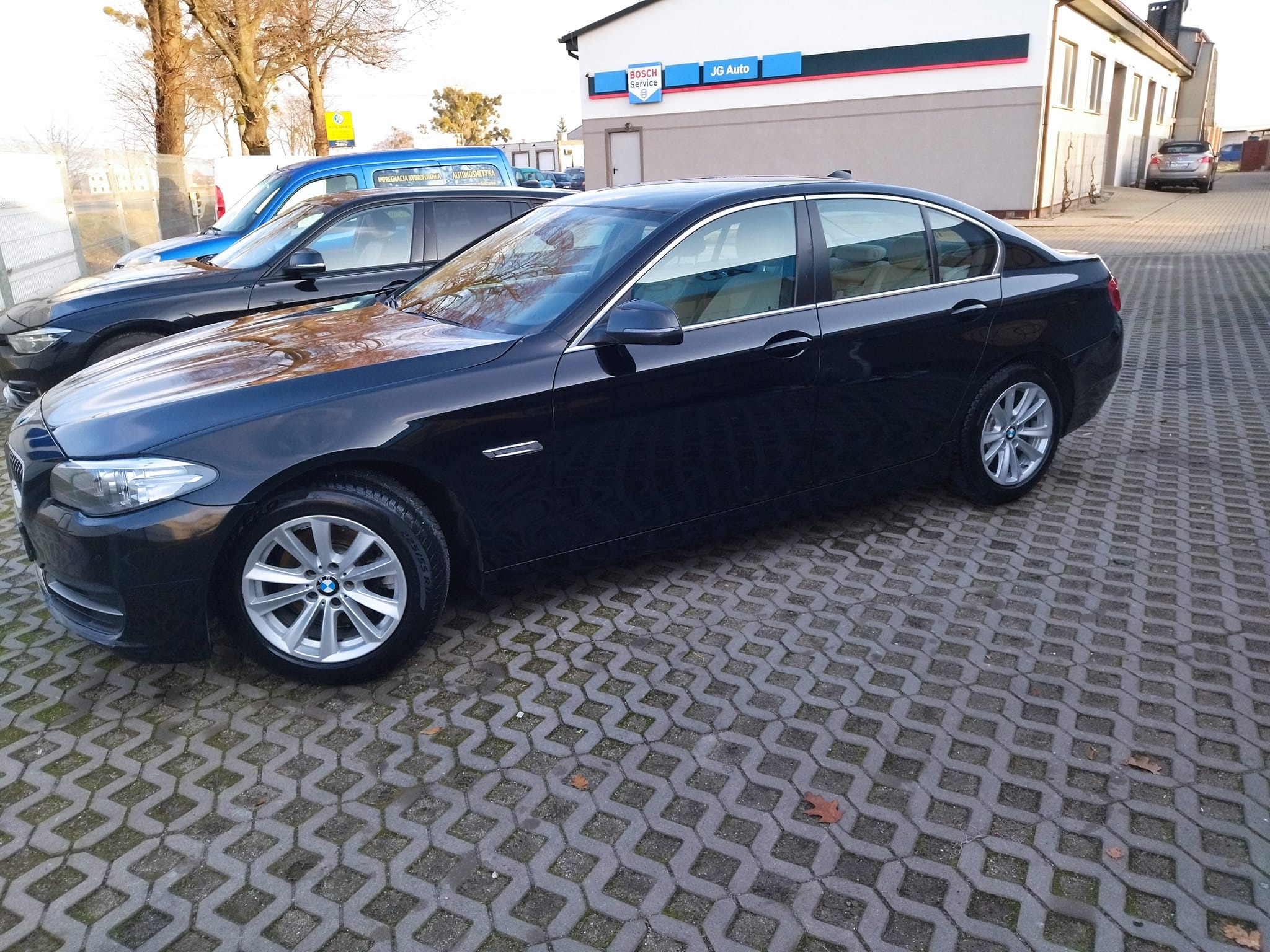 Czarny sedan BMW zaparkowany na kostce brukowej przed budynkiem z logo Bosch Service i JG Auto.