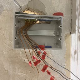 Misha Halka - Otwarta puszka elektryczna zamontowana na surowej, nierównej ścianie z wystającymi przewodami oznaczonymi czerwonymi etykietami.