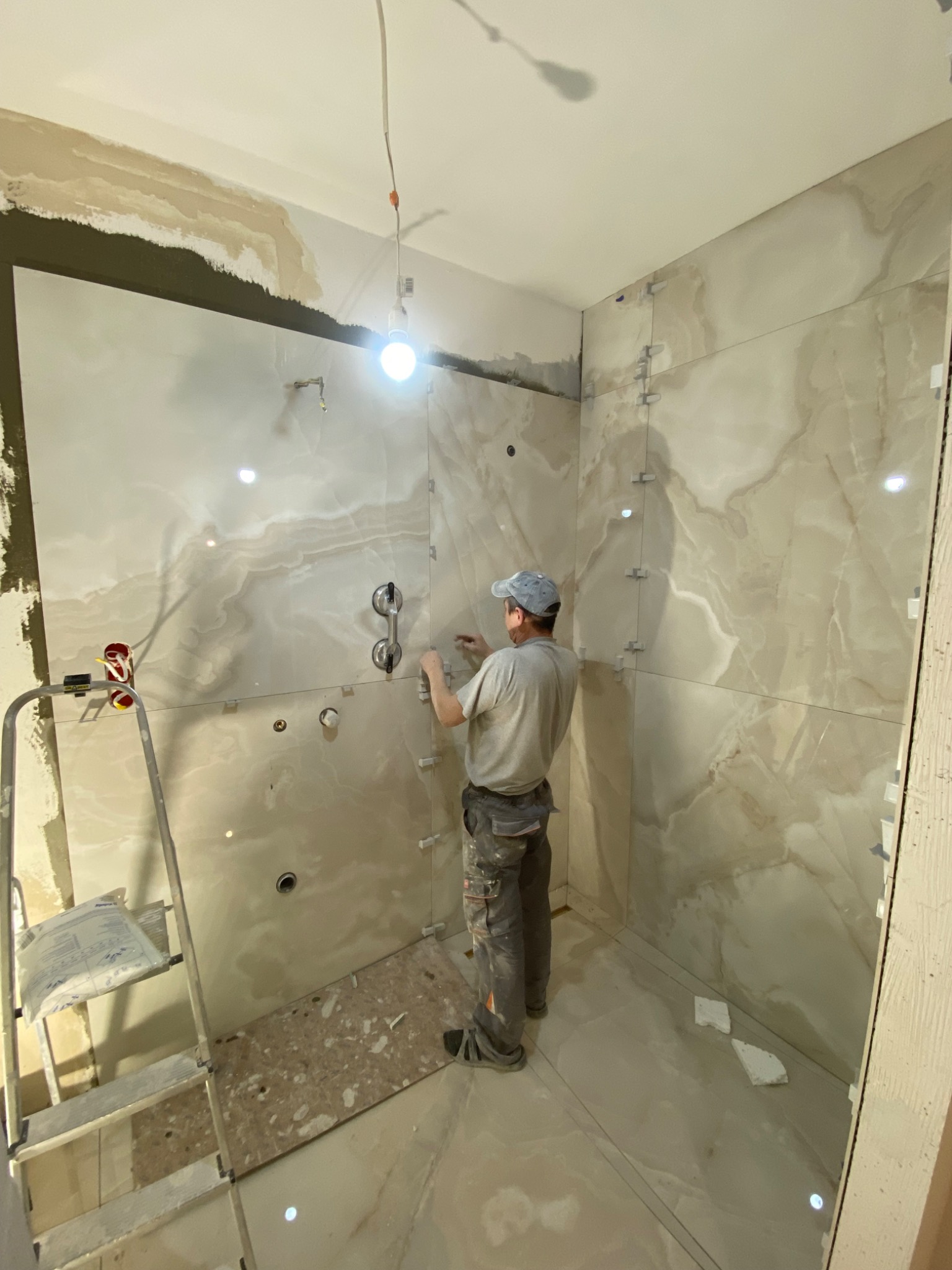 Glazurnik w trakcie montażu płytek imitujących marmur w łazience, używa przyssawek do precyzyjnego umieszczenia płyty, widoczne krzyżyki dystansowe i drabina z workiem zaprawy.