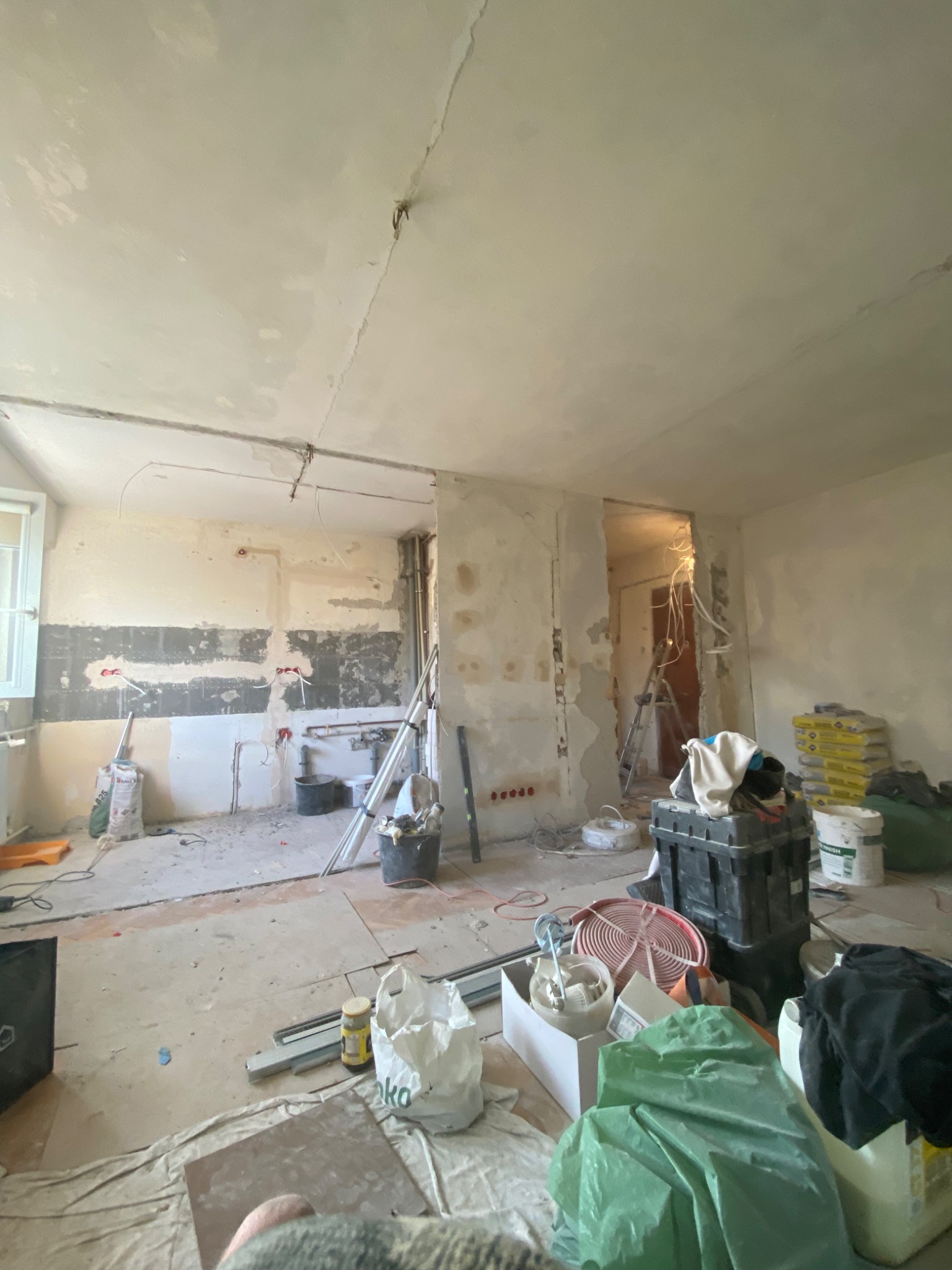 Wnętrze mieszkania w trakcie gruntownego remontu: odsłonięte instalacje elektryczne, częściowo usunięte tynki, widoczne worki z materiałami budowlanymi, drabina i narzędzia rozstawione...