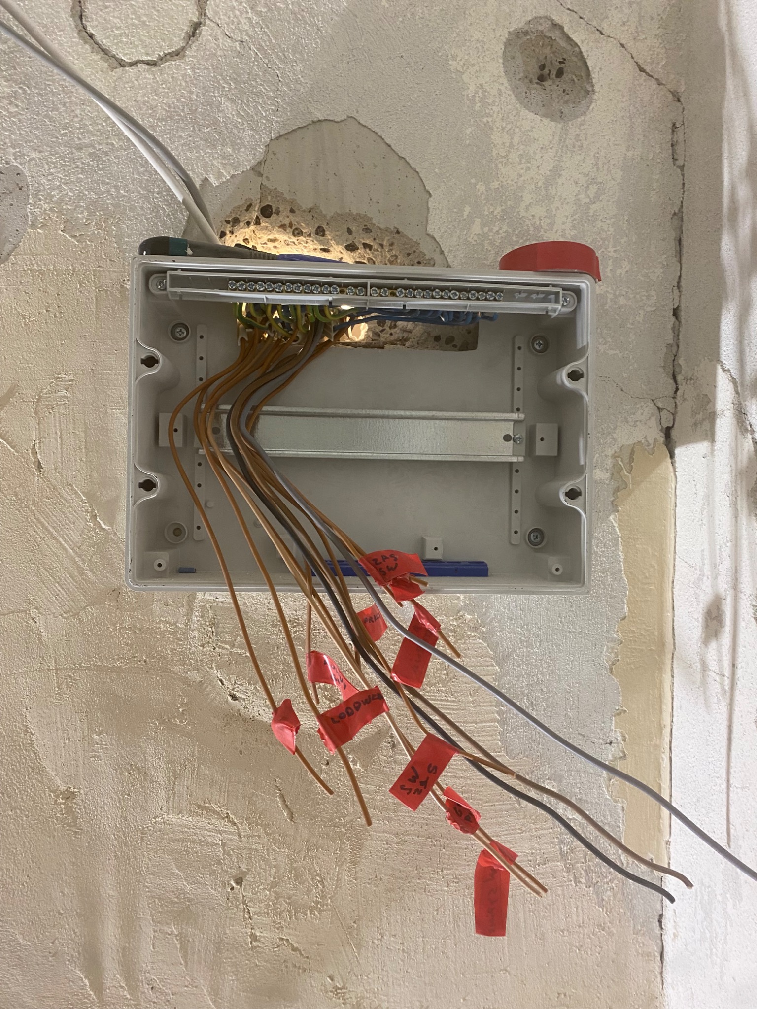 Otwarta puszka elektryczna zamontowana na surowej, nierównej ścianie z wystającymi przewodami oznaczonymi czerwonymi etykietami.