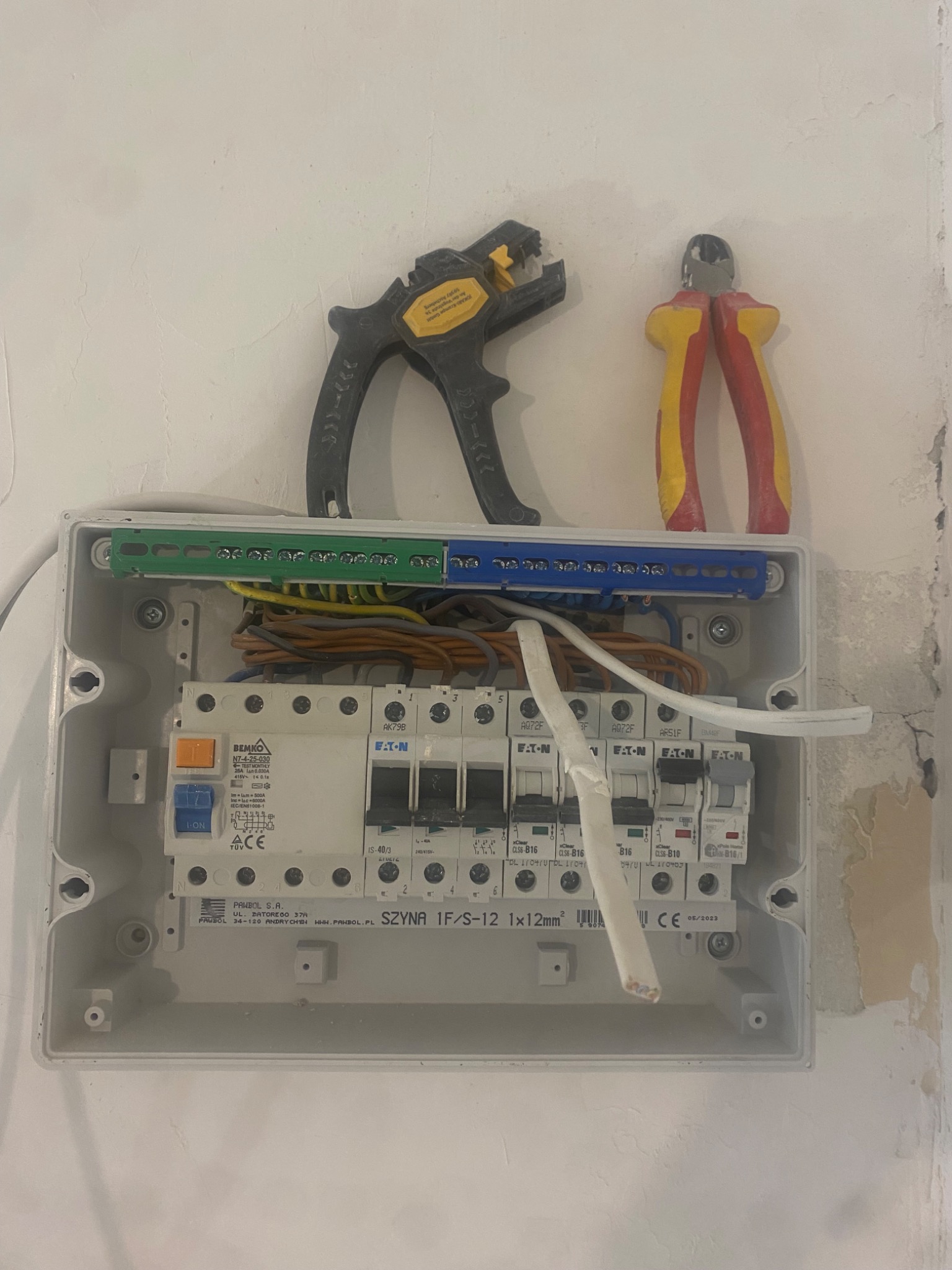 Otwarta skrzynka elektryczna z zainstalowanymi bezpiecznikami Eaton, widoczne przewody, listwy zaciskowe, obok leżą ściągacz izolacji Jokari i obcinaczki boczne z żółto-czerwonymi rękojeściami...