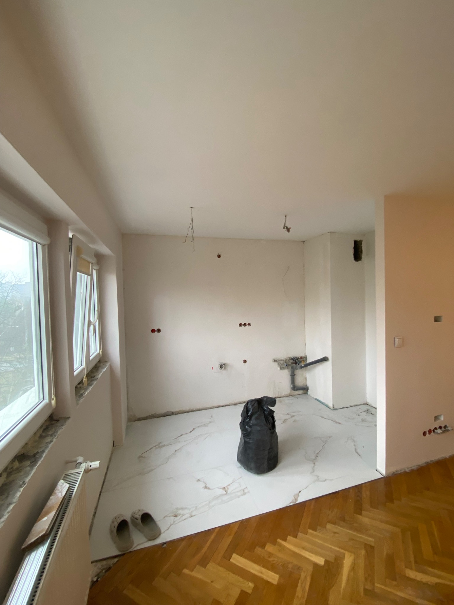 Wnętrze w trakcie prac przygotowawczych do remontu: nowa biała marmurowa podłoga styka się ze starą parkietową, ściany częściowo przygotowane do malowania, widoczne instalacje elektryczne...