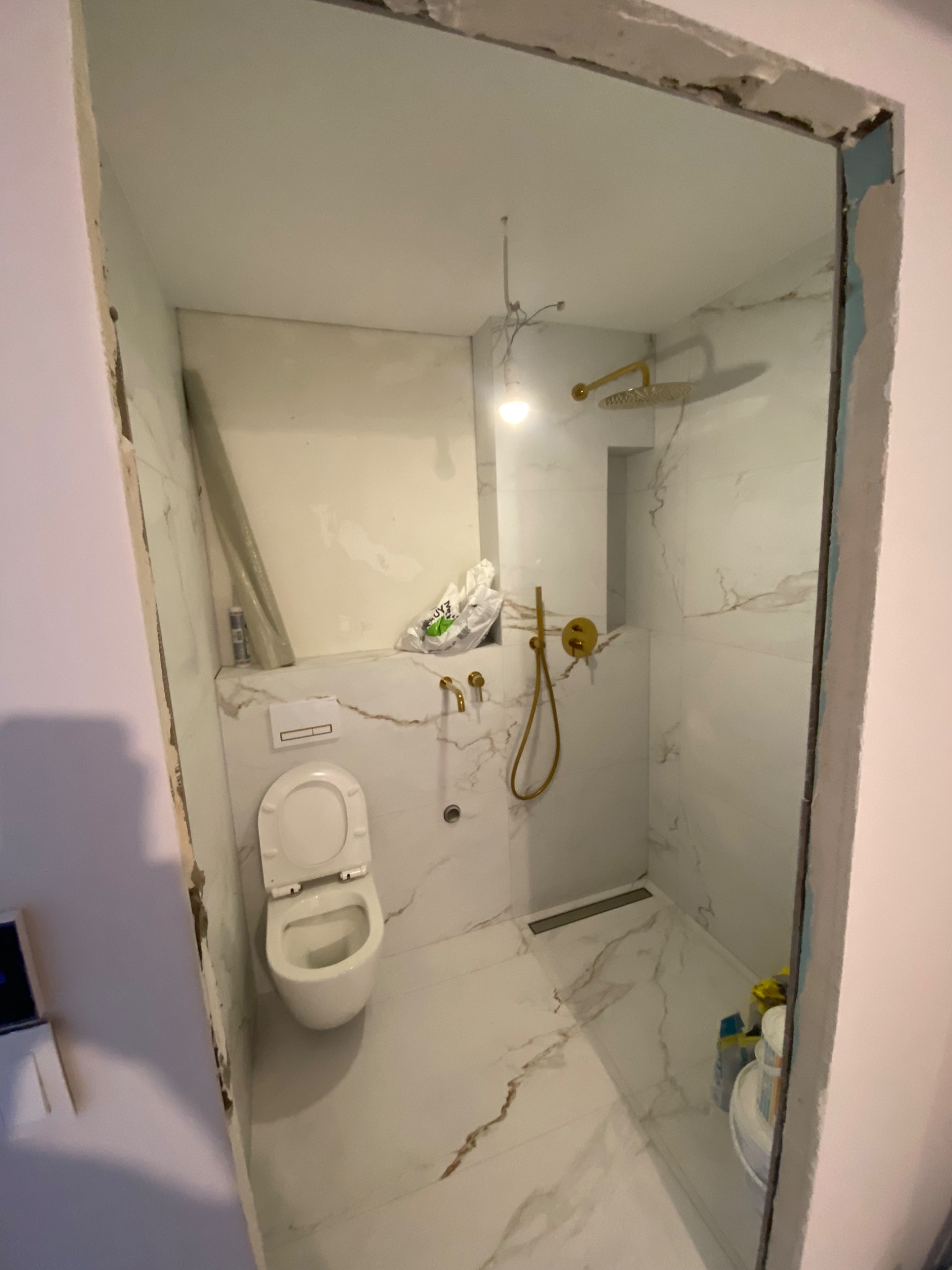Łazienka w trakcie wykańczania: białe marmurowe płytki, złota armatura prysznicowa, podwieszana toaleta, widoczne worki z materiałami budowlanymi i niedokończone obramowanie drzwi.