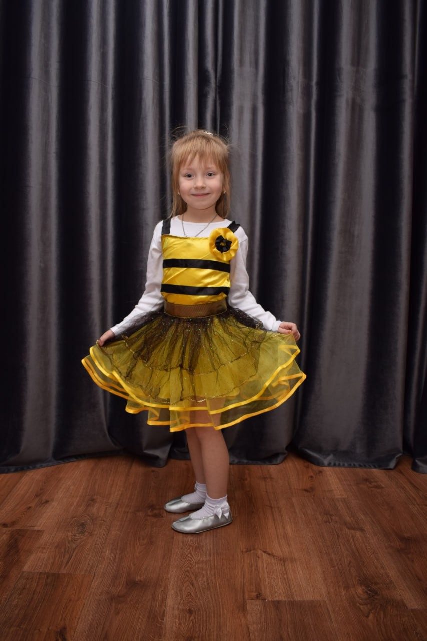 Uśmiechnięta dziewczynka w stroju pszczoły z czarno-żółtym gorsem i tiulową spódnicą, srebrne baletki, na tle szarych zasłon i drewnianej podłogi.