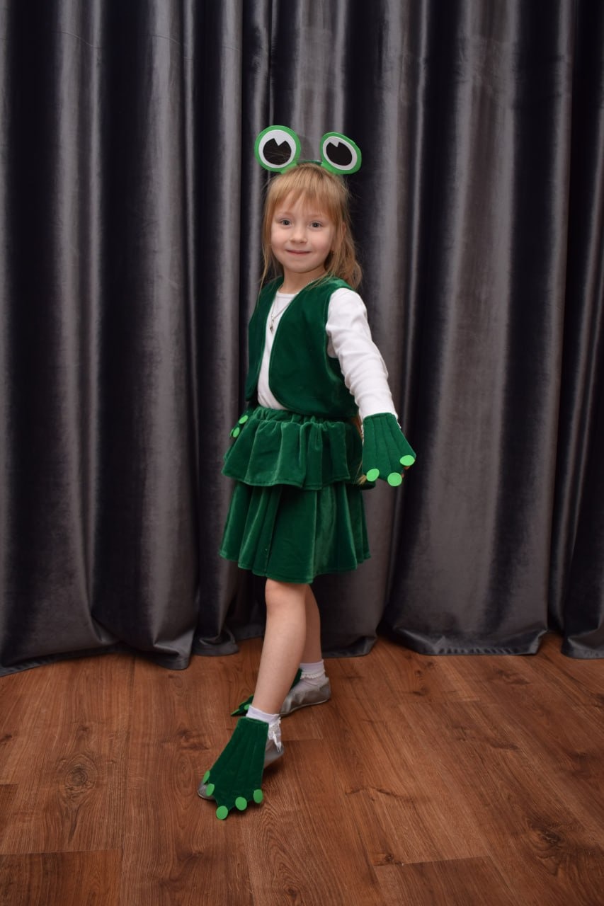 Dziewczynka w zielonym kostiumie żaby z dużymi oczami na opasce, prezentująca strój z Rzeszowa na drewnianej podłodze i tle szarych zasłon.