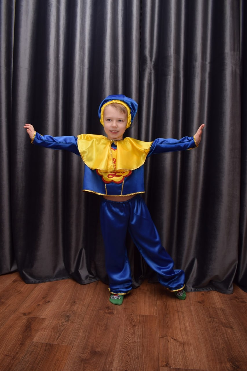 Uśmiechnięty chłopiec w niebiesko-żółtym kostiumie z peleryną i czapką, pozuje na tle szarych zasłon i drewnianej podłogi.