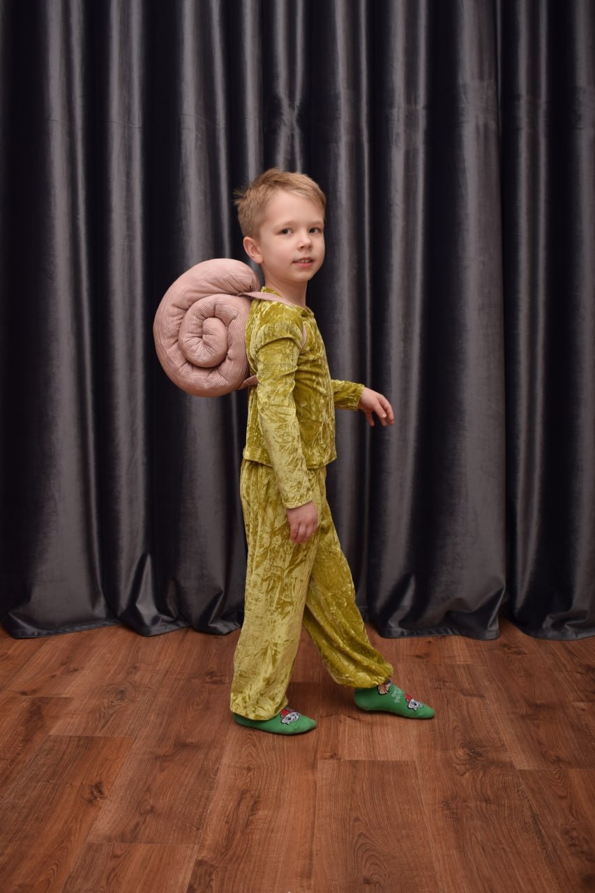 Chłopiec w welurowym kostiumie ślimaka z pluszową muszlą na plecach, stojący na drewnianej podłodze na tle ciemnoszarej zasłony.