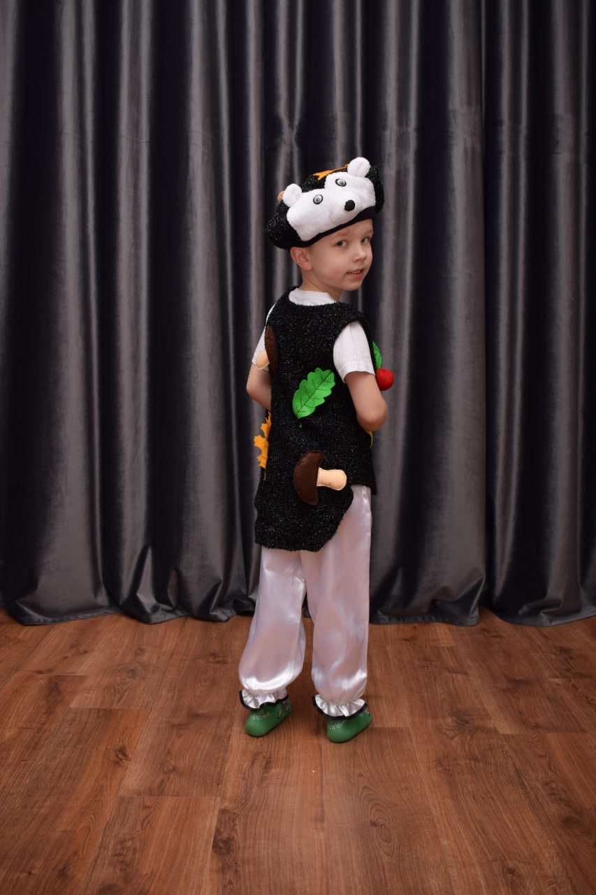 Chłopiec w kostiumie leśnego duszka z czarną kamizelką ozdobioną filcowymi grzybami, liśćmi i jarzębiną, oraz czapką z podobizną zwierzątka, pozuje na tle szarych zasłon.