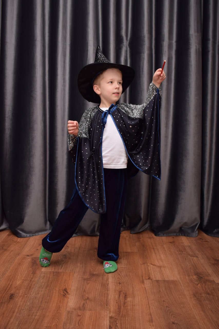 Chłopiec w stroju czarodzieja z różdżką, ubrany w czarny kapelusz i pelerynę w gwiazdki, na tle szarej zasłony i drewnianej podłogi, z zielonymi skarpetkami w łapki.