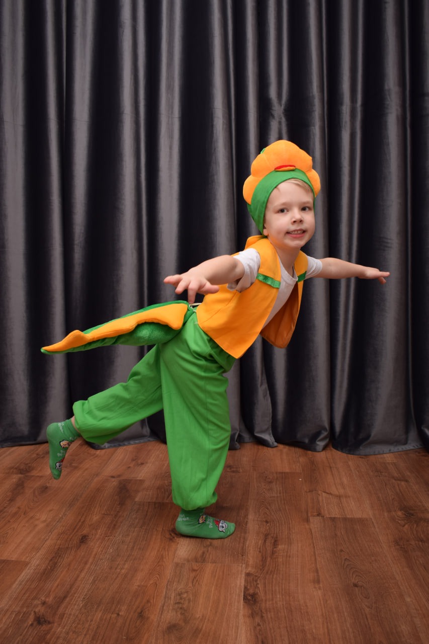 Uśmiechnięty chłopiec w zielono-pomarańczowym stroju warzywa, prawdopodobnie dyni, pozuje na tle szarych zasłon i drewnianej podłogi, stojąc na jednej nodze.
