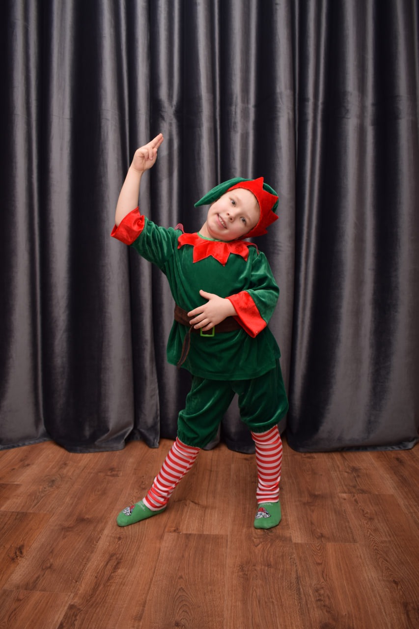 Uśmiechnięty chłopiec w zielono-czerwonym stroju elfa z uniesioną ręką na tle szarej zasłony i drewnianej podłogi.