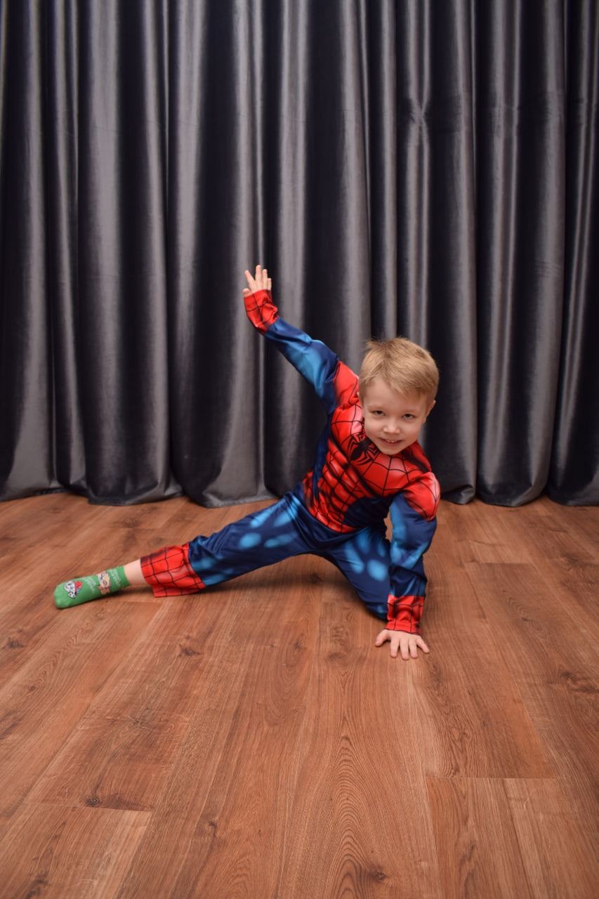 Uśmiechnięty chłopiec w stroju Spider-Mana, pozujący na drewnianej podłodze przed szarymi zasłonami, z zieloną skarpetką w świąteczne wzory na jednej nodze.