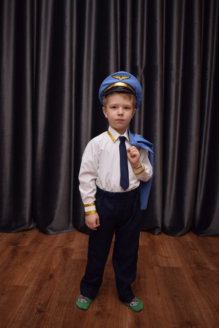 Chłopiec w stroju pilota z kurtką przewieszoną przez ramię, pozuje na tle szarych zasłon i drewnianej podłogi, w zielonych kapciach z motywem z bajki.