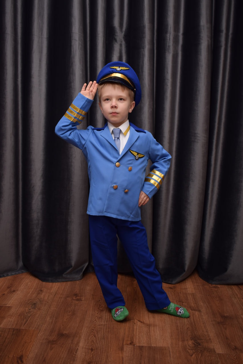 Chłopiec w kostiumie pilota, salutujący, z zielonymi skarpetkami w kreskówkowe wzory, na tle szarych zasłon i drewnianej podłogi.
