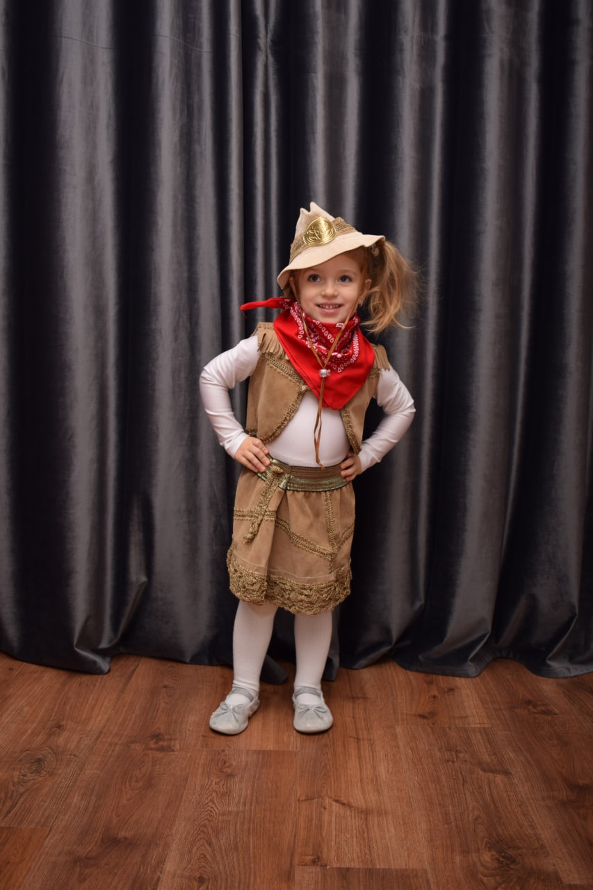 Uśmiechnięta dziewczynka w stroju kowbojki, z czerwonym bandaną i kapeluszem z ozdobą, pozuje na tle szarej zasłony.
