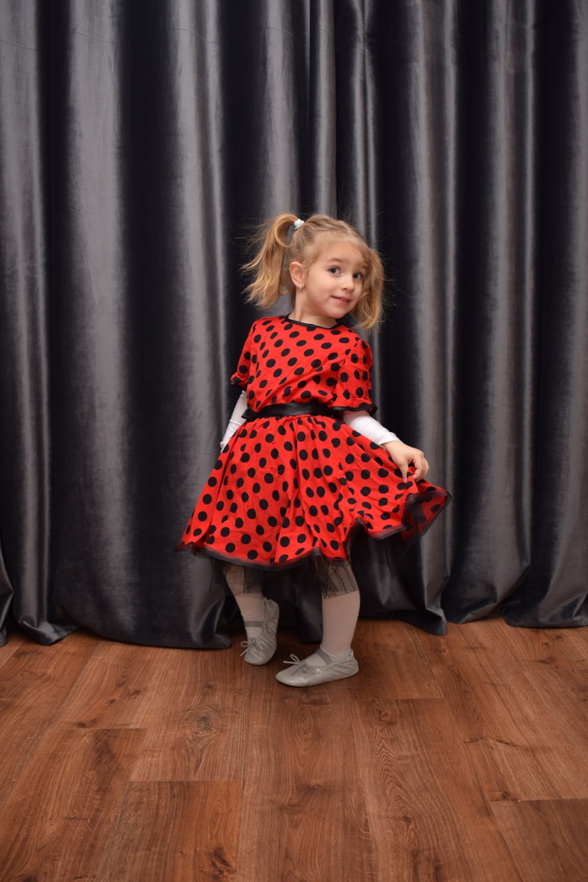 Dziewczynka w czerwonej sukience w czarne kropki, z czarną tiulową podszewką i baletkami, pozuje na tle szarej, drapowanej zasłony.