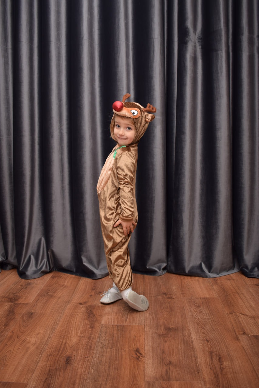 Uśmiechnięta dziewczynka w kostiumie renifera z czerwonym nosem, pozująca na tle szarych zasłon i drewnianej podłogi.