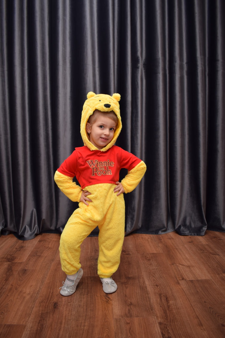 Uśmiechnięta dziewczynka w kostiumie Kubusia Puchatka z kapturem z uszami, pozująca na tle szarych zasłon i drewnianej podłogi, z rękami na biodrach i srebrnymi baletkami na stopach.
