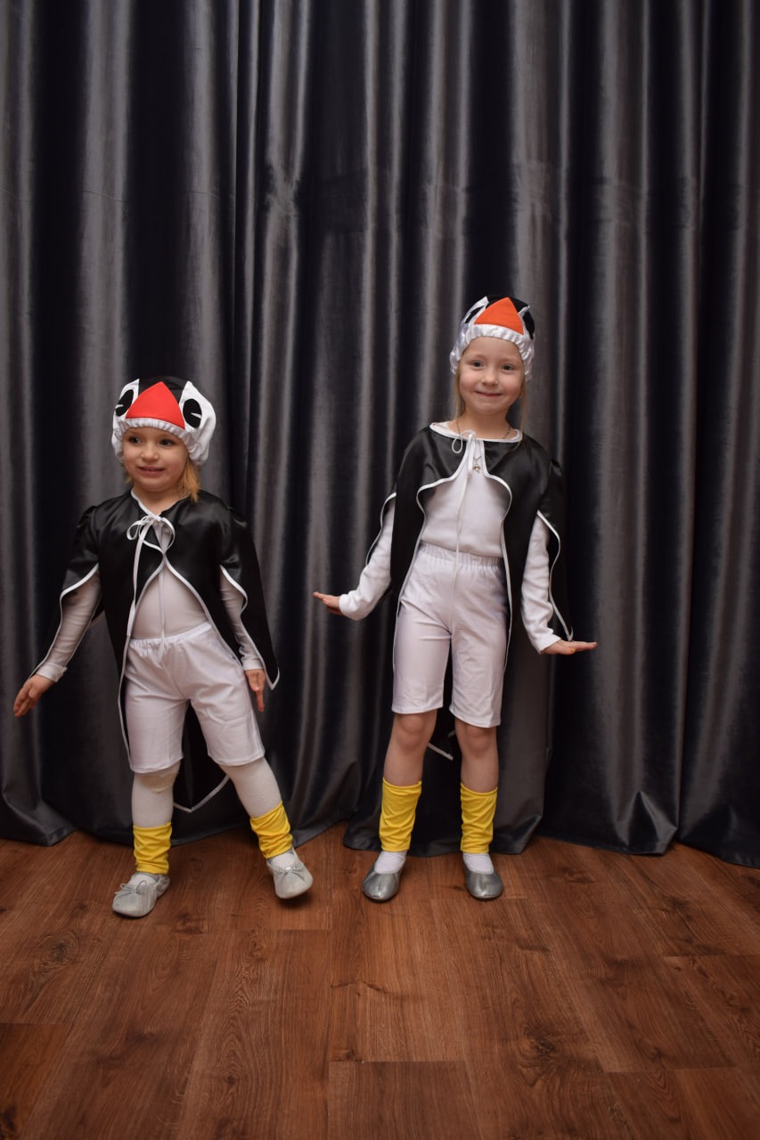 Dwie uśmiechnięte dziewczynki w kostiumach pingwinów z czarnymi pelerynami i białymi strojami, stojące na drewnianej podłodze przed ciemnoszarymi zasłonami.