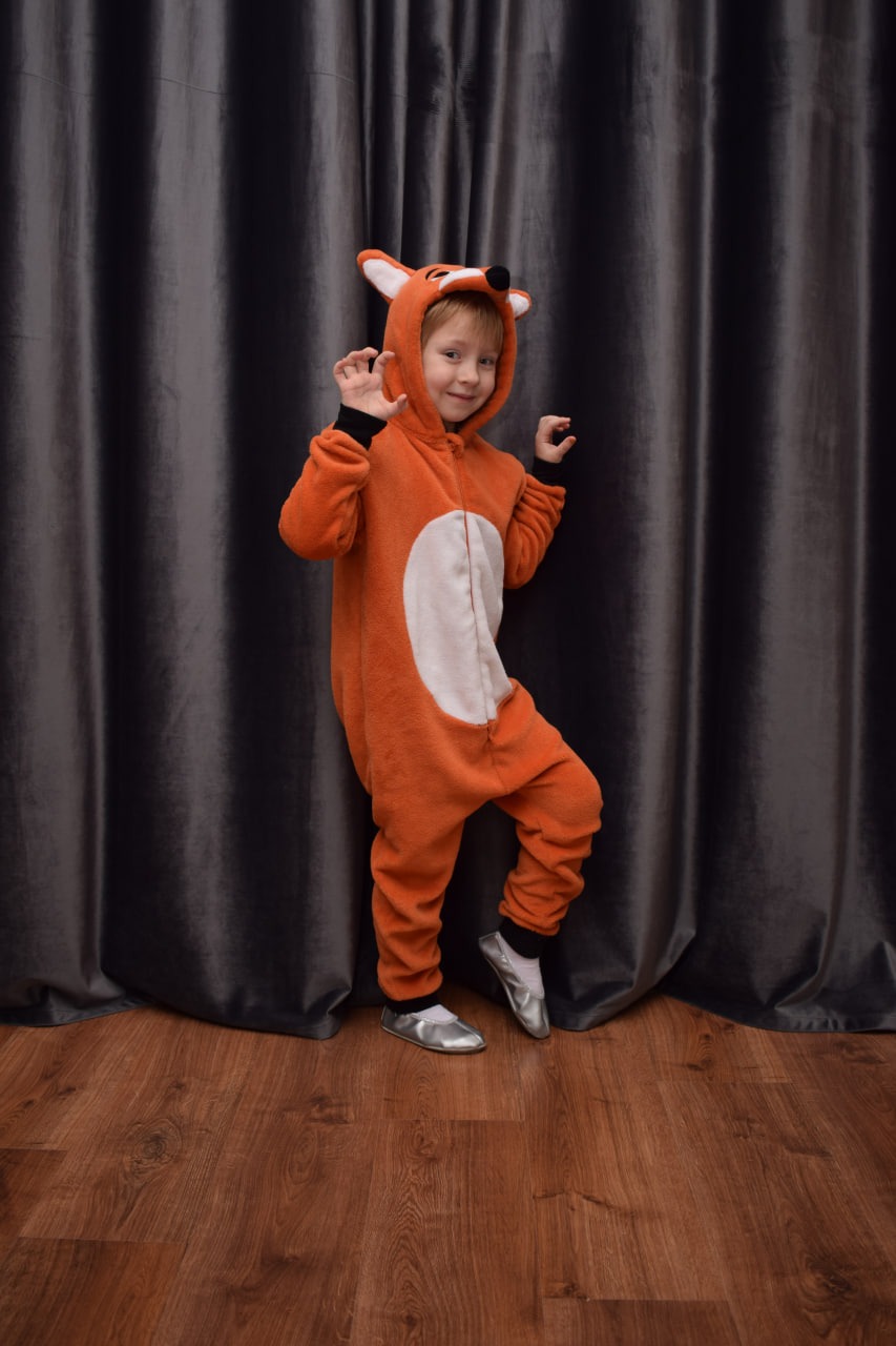 Chłopiec w pomarańczowym kombinezonie lisa z kapturem, pozujący na tle ciemnoszarej zasłony i drewnianej podłogi.